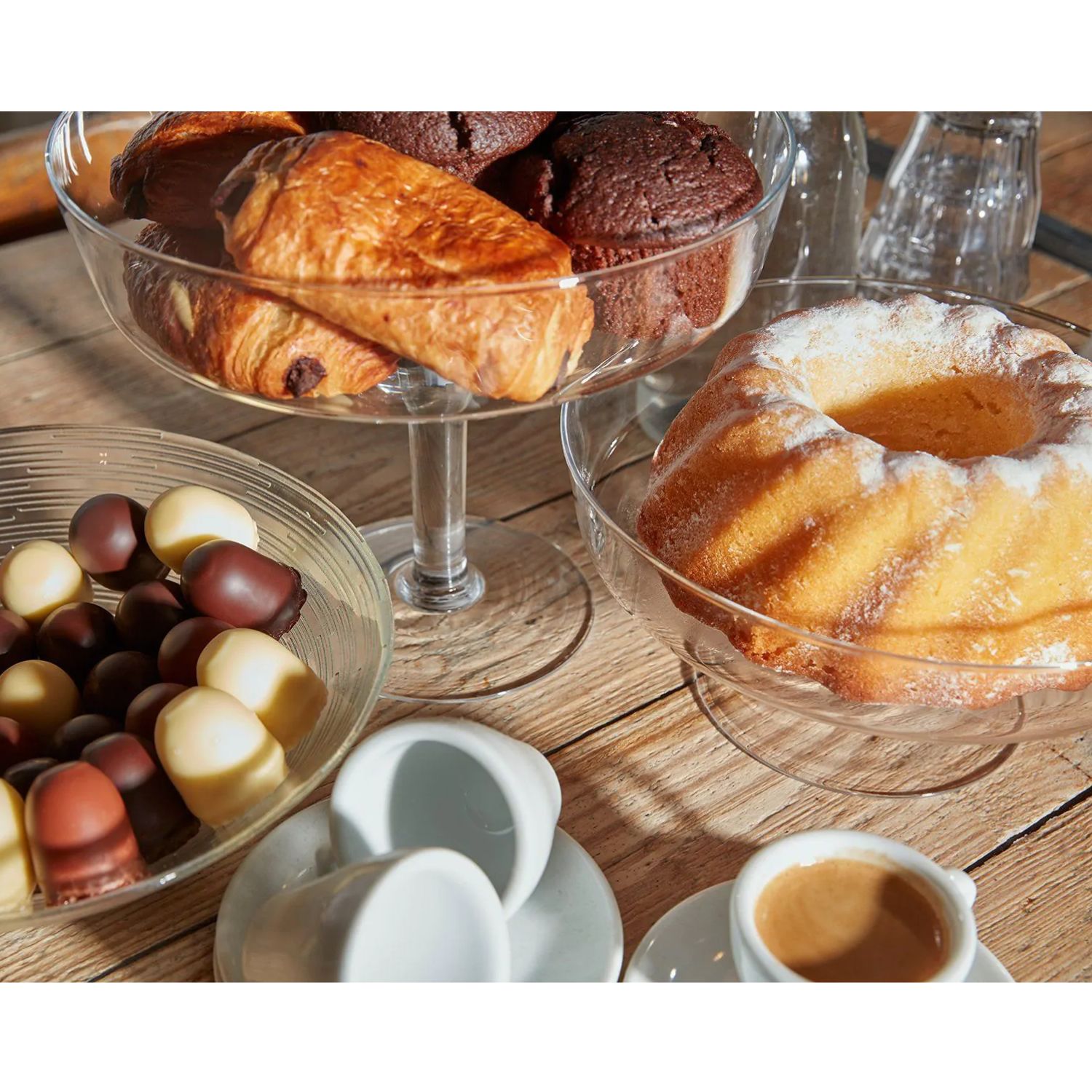 Gugelhupf, Croissants und Muffins auf Glasschalen, Pralinen und Espresso auf Holztisch