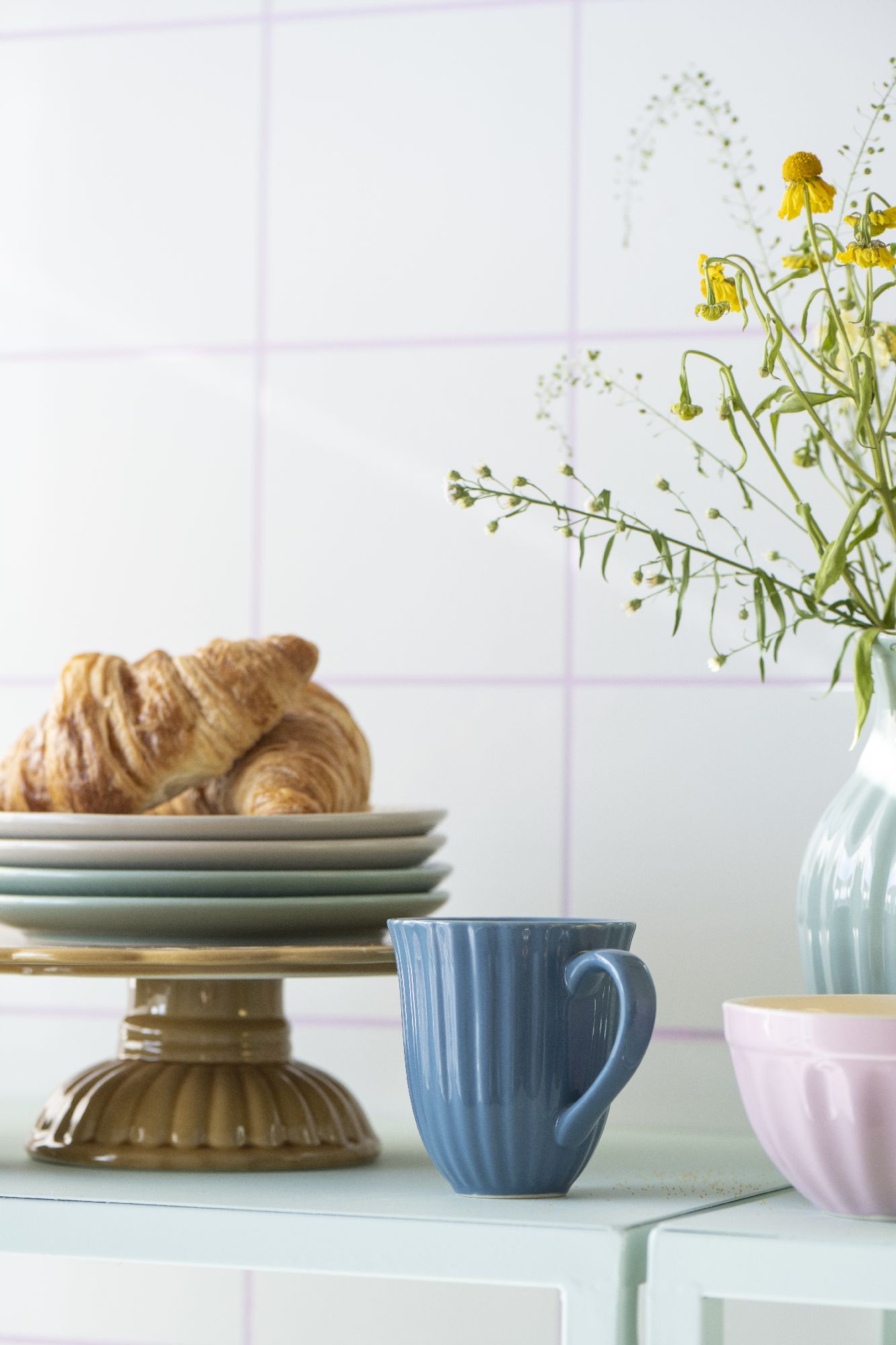 Ein blauer Becher steht neben einem Teller mit Croissants und mehreren Schalen auf einem Tisch. Im Hintergrund sind Blumen in einer Vase.