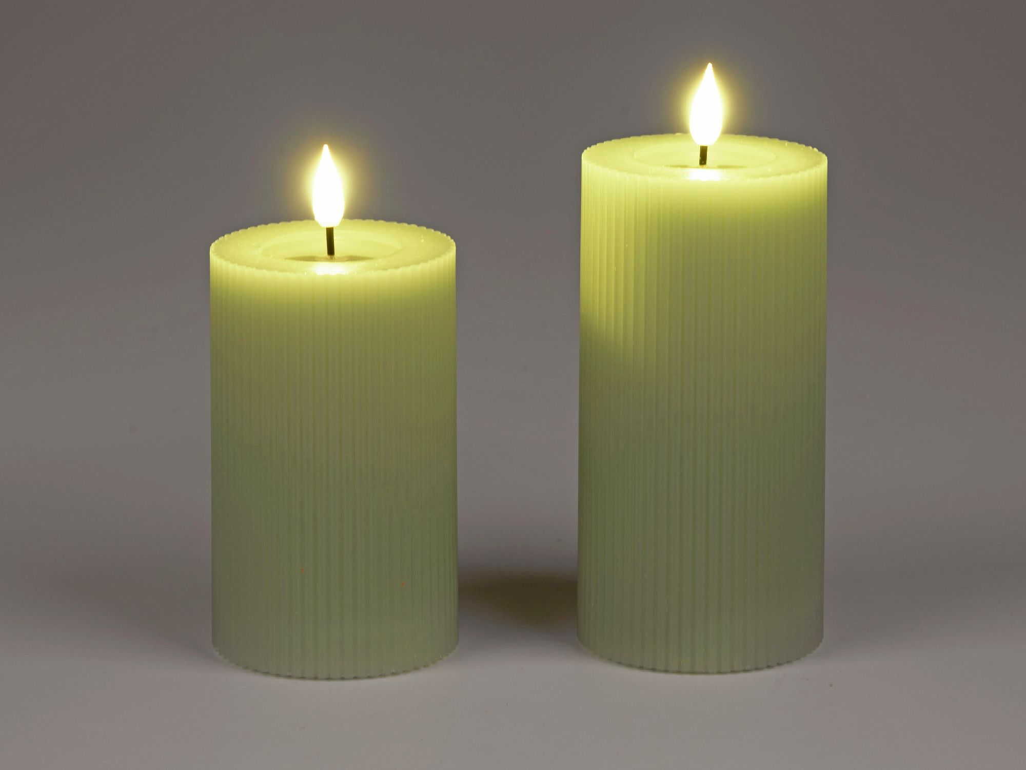 Zwei grüne, gerippte Kerzen mit flackernden Flammen stehen nebeneinander auf einem neutralen Hintergrund.