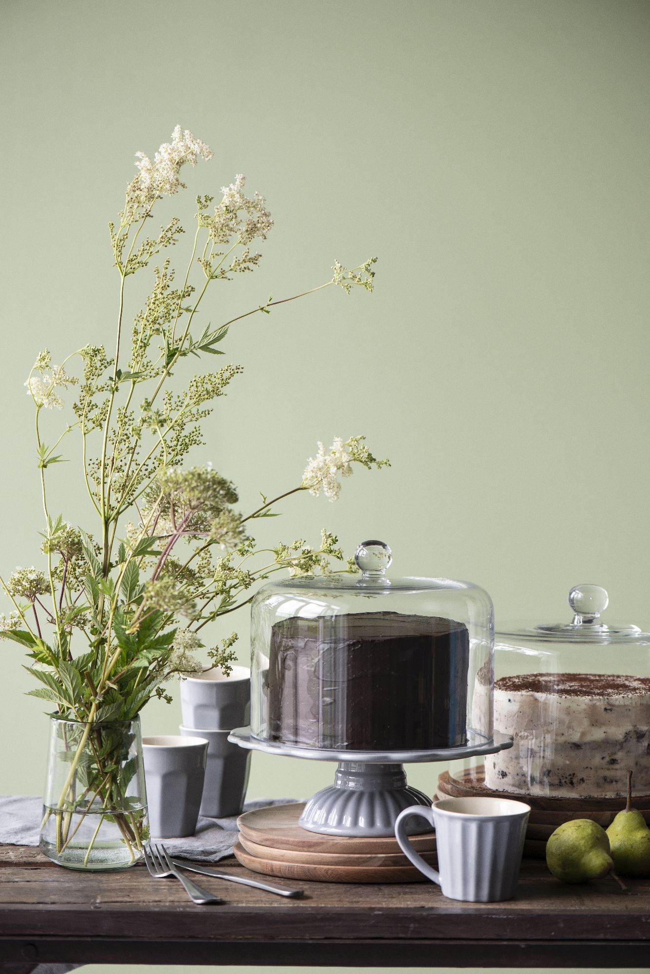 Ein Tisch mit zwei Torten unter Glasglocken, einer Vase mit Blumen, grauen Tassen und einer Birne auf einem Holzbrett.