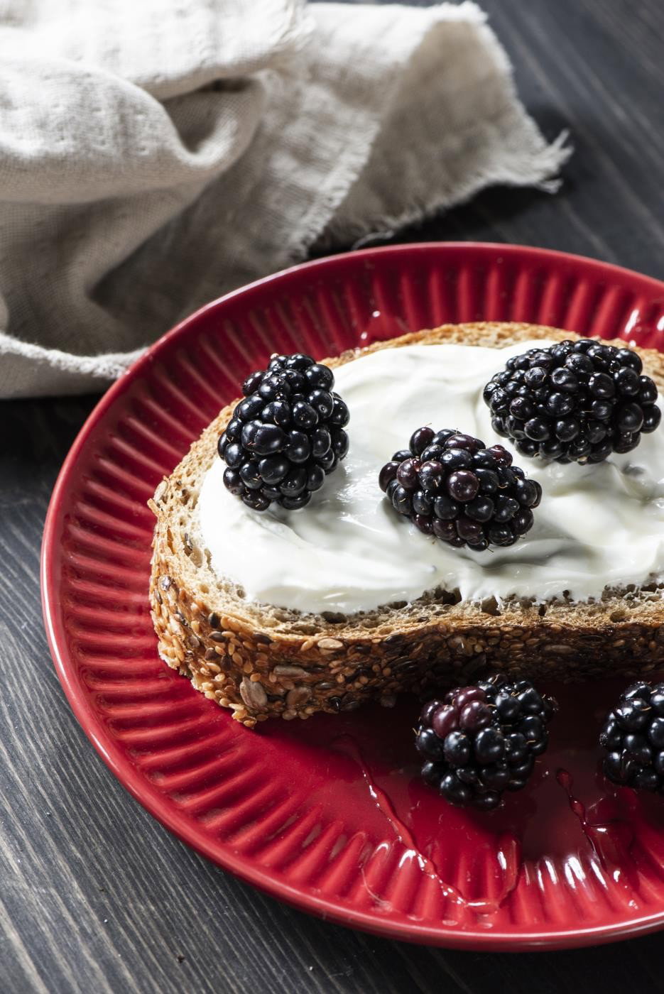 Ein Stück Vollkornbrot mit Joghurt und frischen Brombeeren auf einem roten Teller, daneben ein graues Tuch.