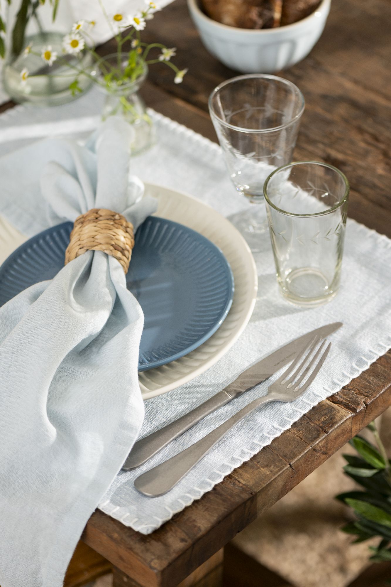 Tischgedeck mit blauer und weißer Teller, Besteck, Gläsern und einer Serviette, auf einem Holzuntergrund. Blumen in der Nähe.