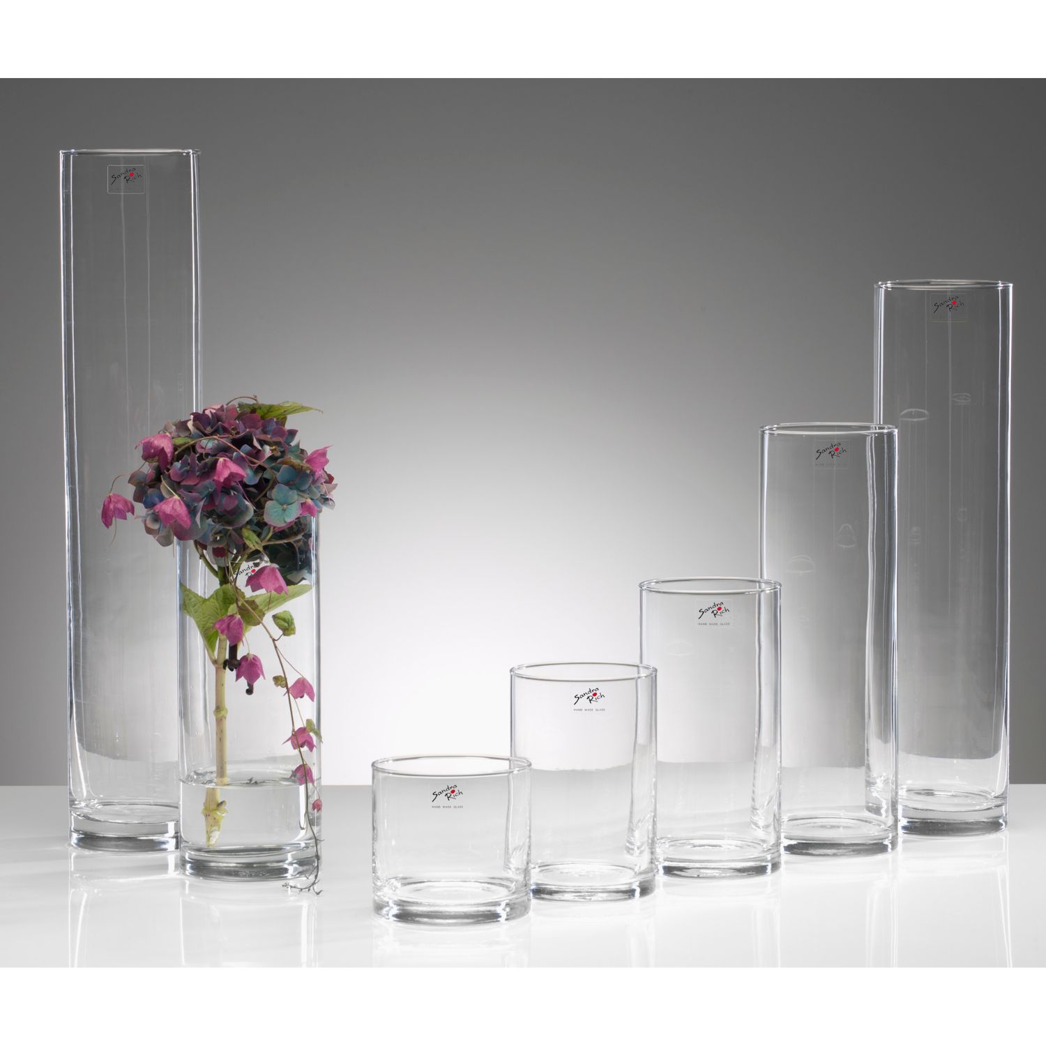 Sechs klare Glasvasen in verschiedenen Höhen, links eine mit lila Blumen