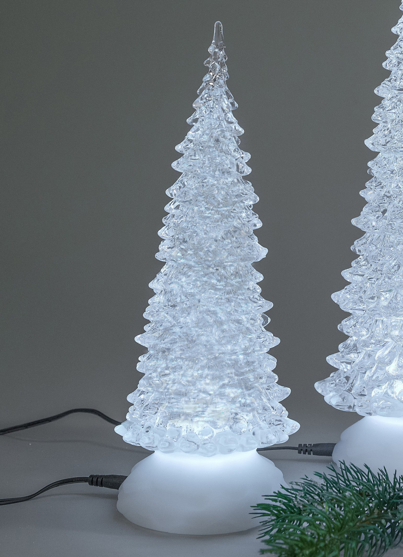 Zwei leuchtende, gläserne Weihnachtsbaumfiguren stehen auf einem weißen Sockel, umgeben von einem gr