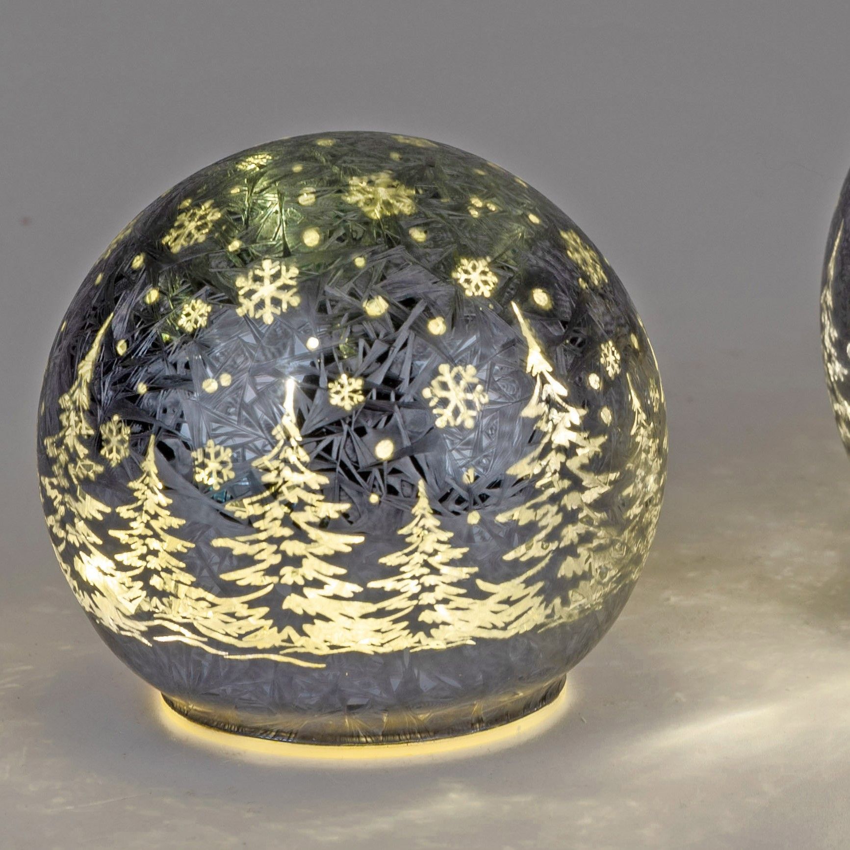 Eine gläserne Kugel mit winterlichem Motiv: weiße Tannen und Schneeflocken auf dunklem Hintergrund,