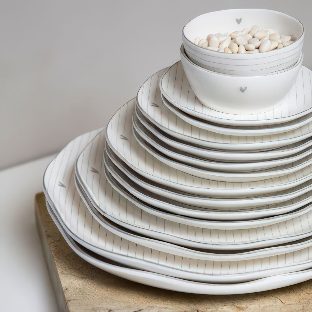 Gestapelte, weiße Teller mit feinen Linien und ein kleinerer Schüssel mit weißen Bohnen in der Mitte auf einem Holzbrett.