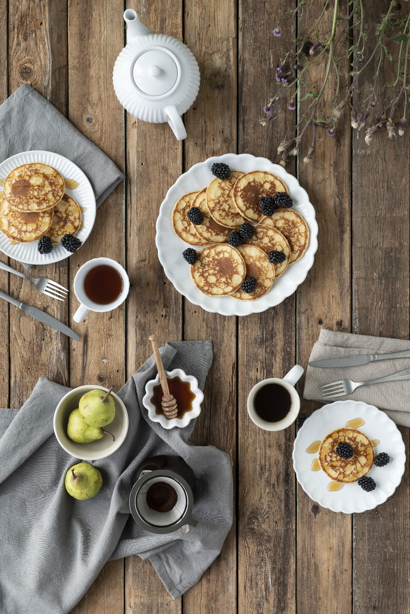 Ein Tisch mit Pfannkuchen, Brombeeren, Tee, Kaffee, Birnen und Honig auf einem grauen Tuch, umgeben von Holzoberfläche.