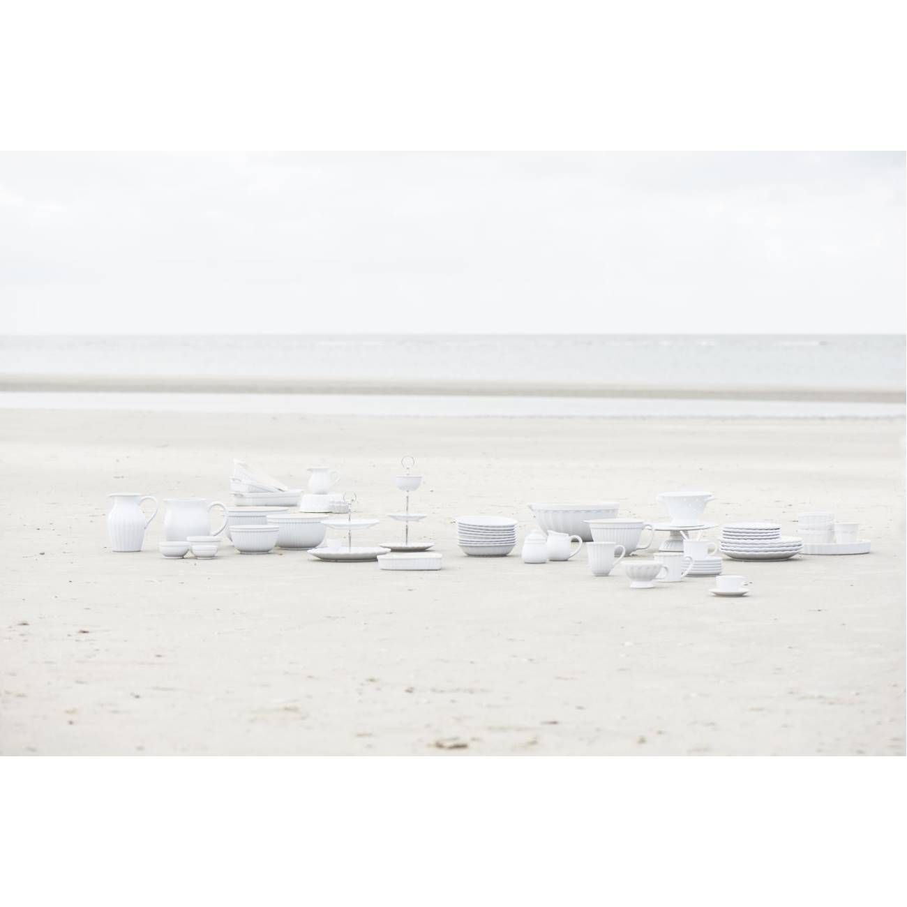 Eine Ansammlung von weißen Geschirrteilen auf einem sandigen Strand, mit dem Meer im Hintergrund und bewölktem Himmel.
