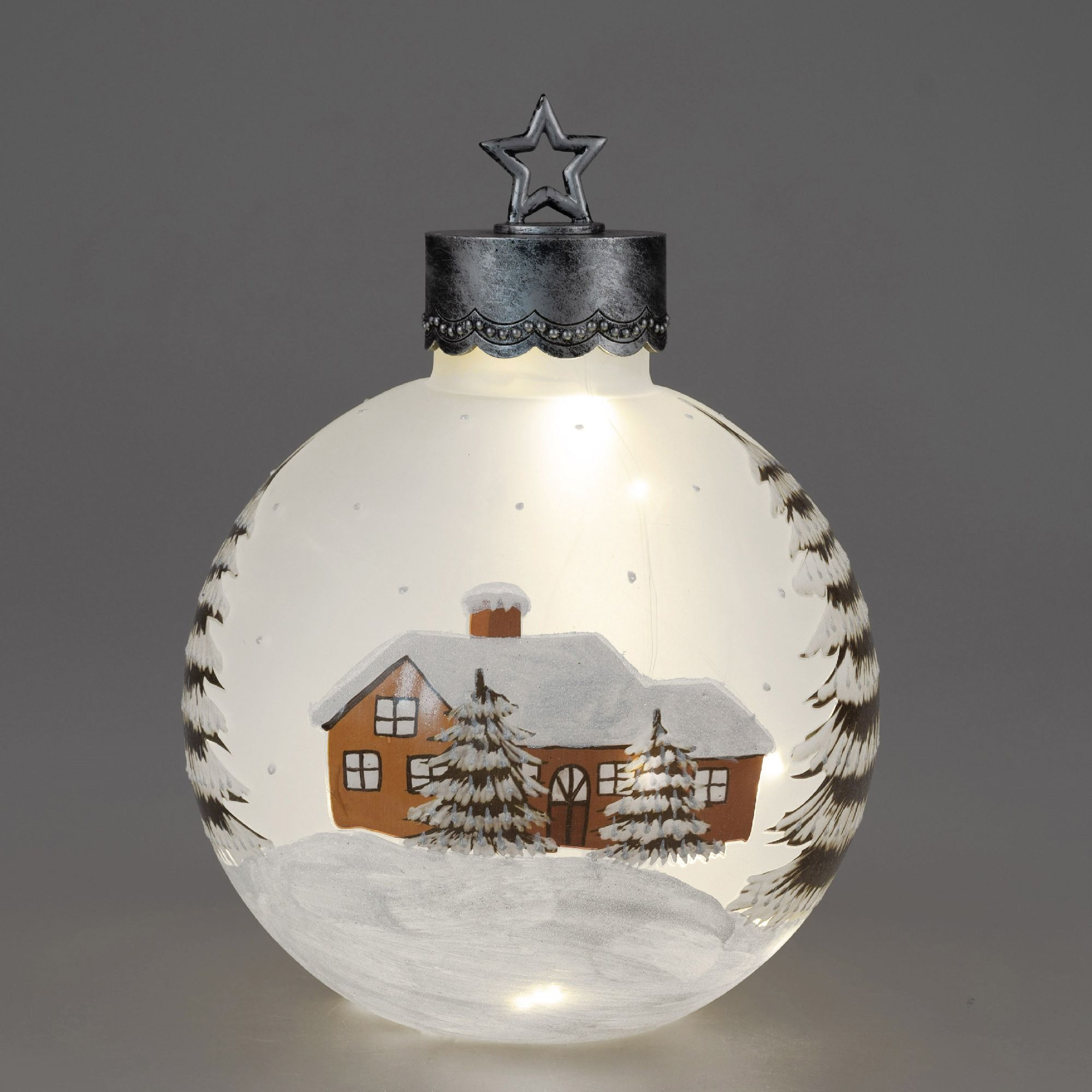 Eine gläserne Weihnachtskugel mit einem bemalten Haus und Tannenbäumen in einer Schneelandschaft. Ob