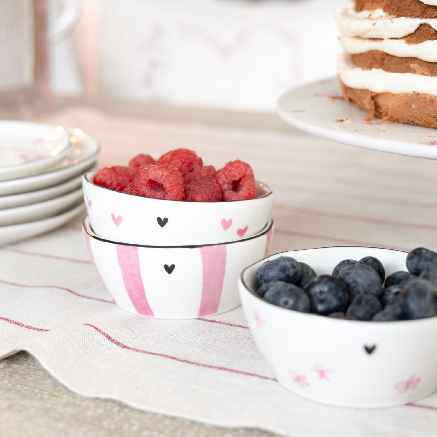 Schalen mit Himbeeren und Blaubeeren auf gestreifter Tischdecke, daneben ein angeschnittener Kuchen