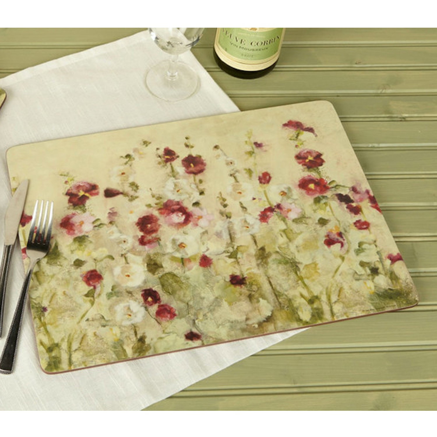 Ein Tischset mit floralen Motiven in sanften Farben liegt auf einem hellen Tisch. Besteck und ein Glas sind daneben platziert.