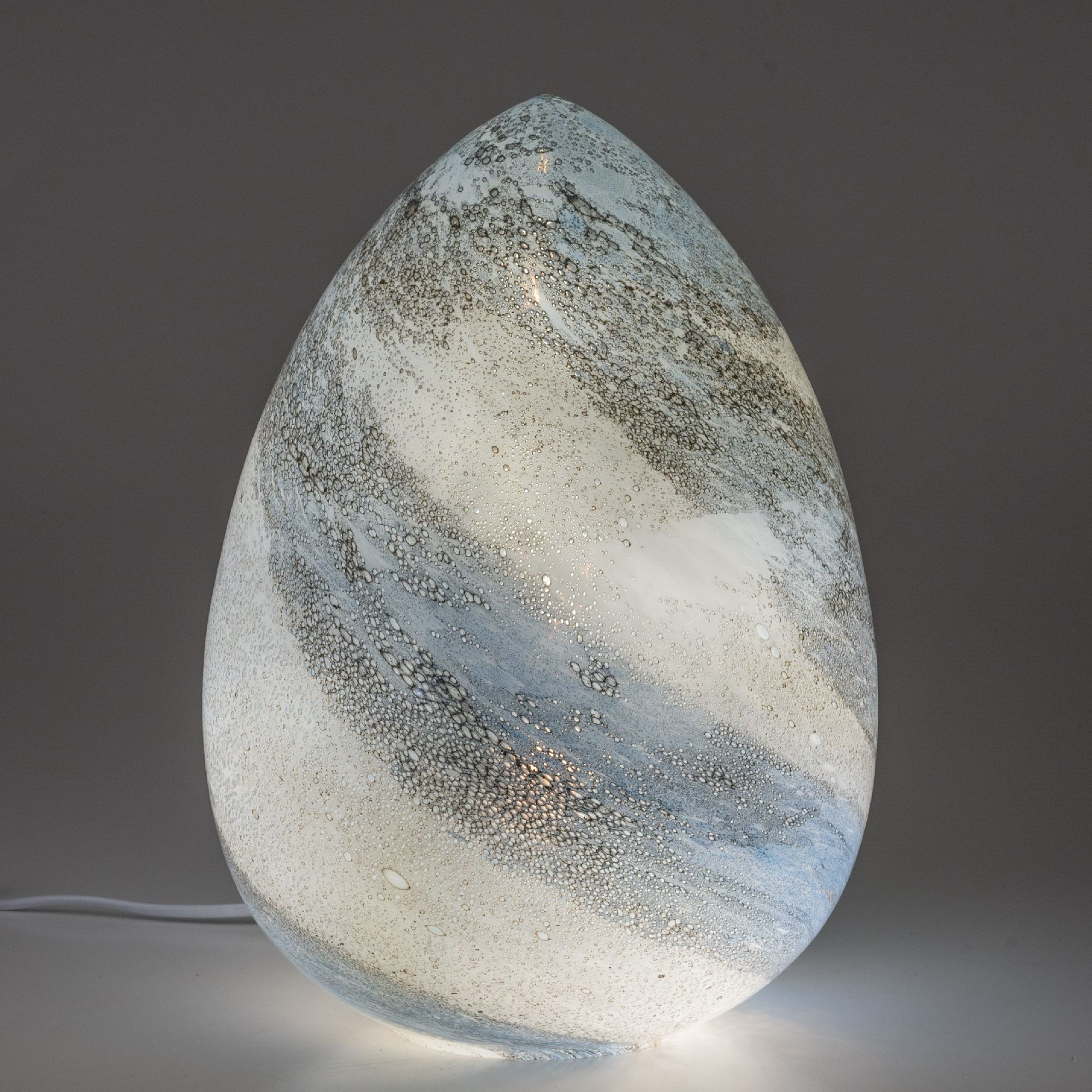 Eine gläserne Lampe in Eiform mit blauen und weißen Farbverläufen sowie kleinen Blasen, die sanft leuchtet.