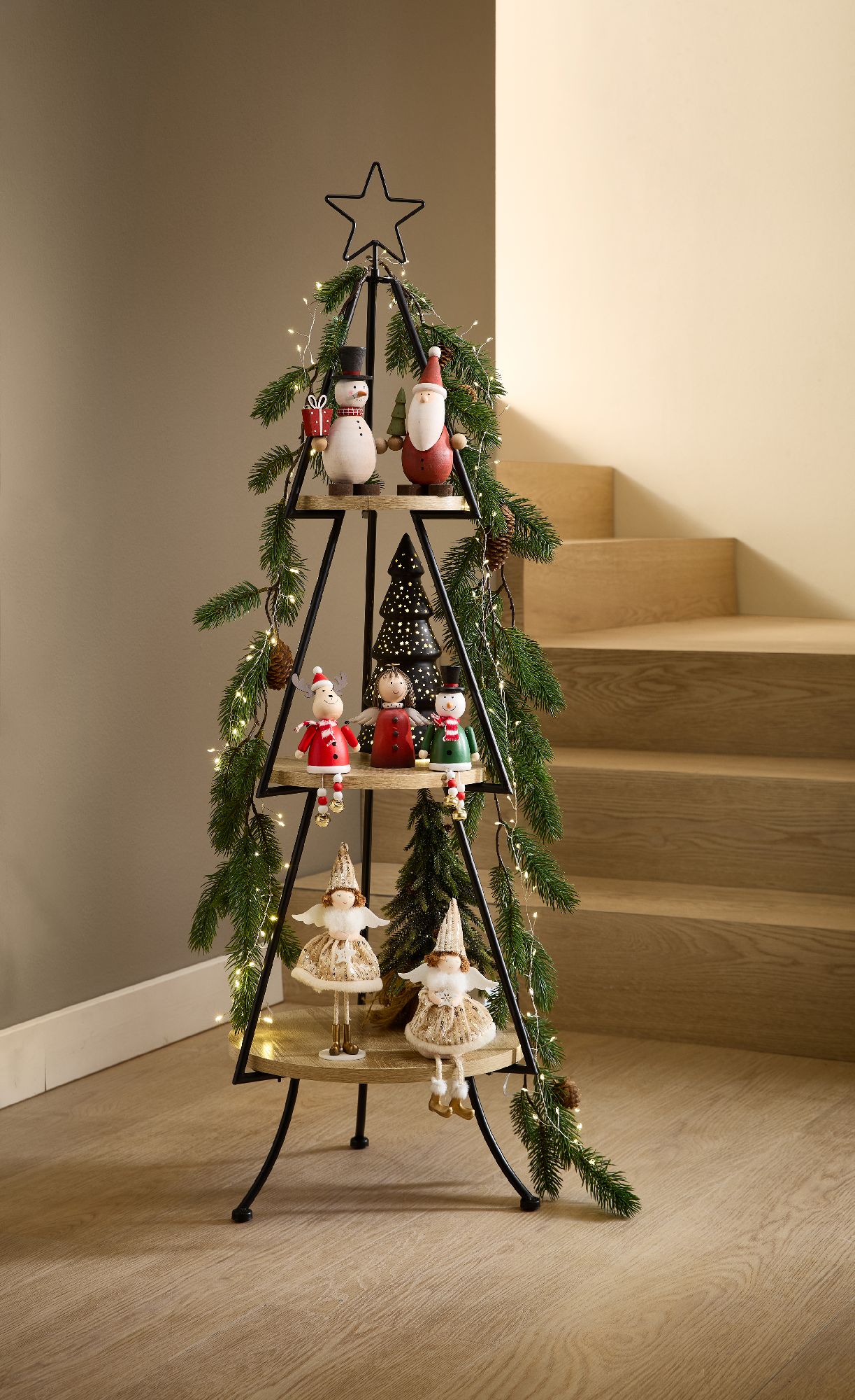 Dekorativer Weihnachtsbaum aus Metall mit Figuren, Lichtern und Tannenzweigen, vor einer Treppe auf einem Holzfußboden.