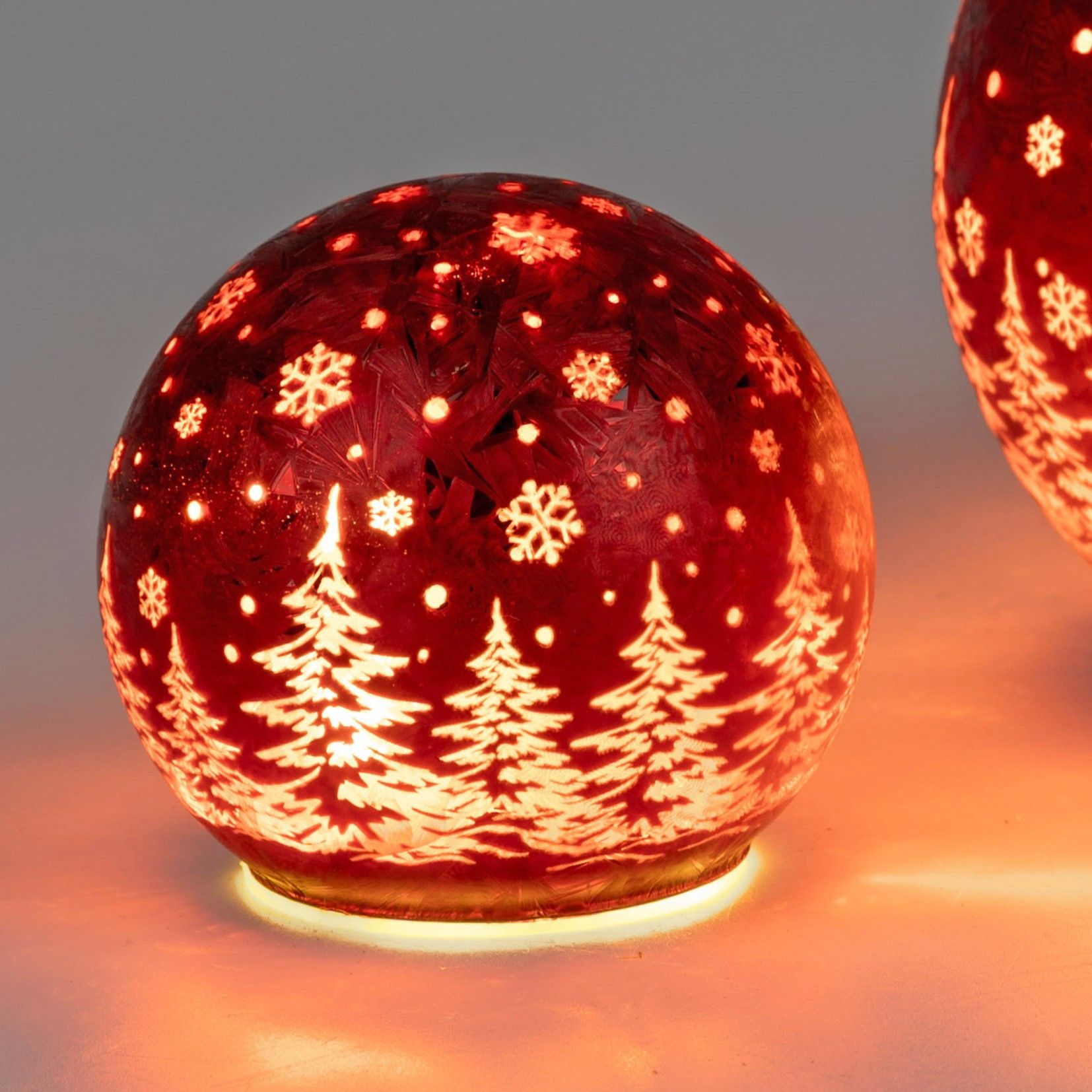 Rote Weihnachtskugel mit weißen Tannenbäumen und Schneeflocken, beleuchtet von innen, auf einem hell