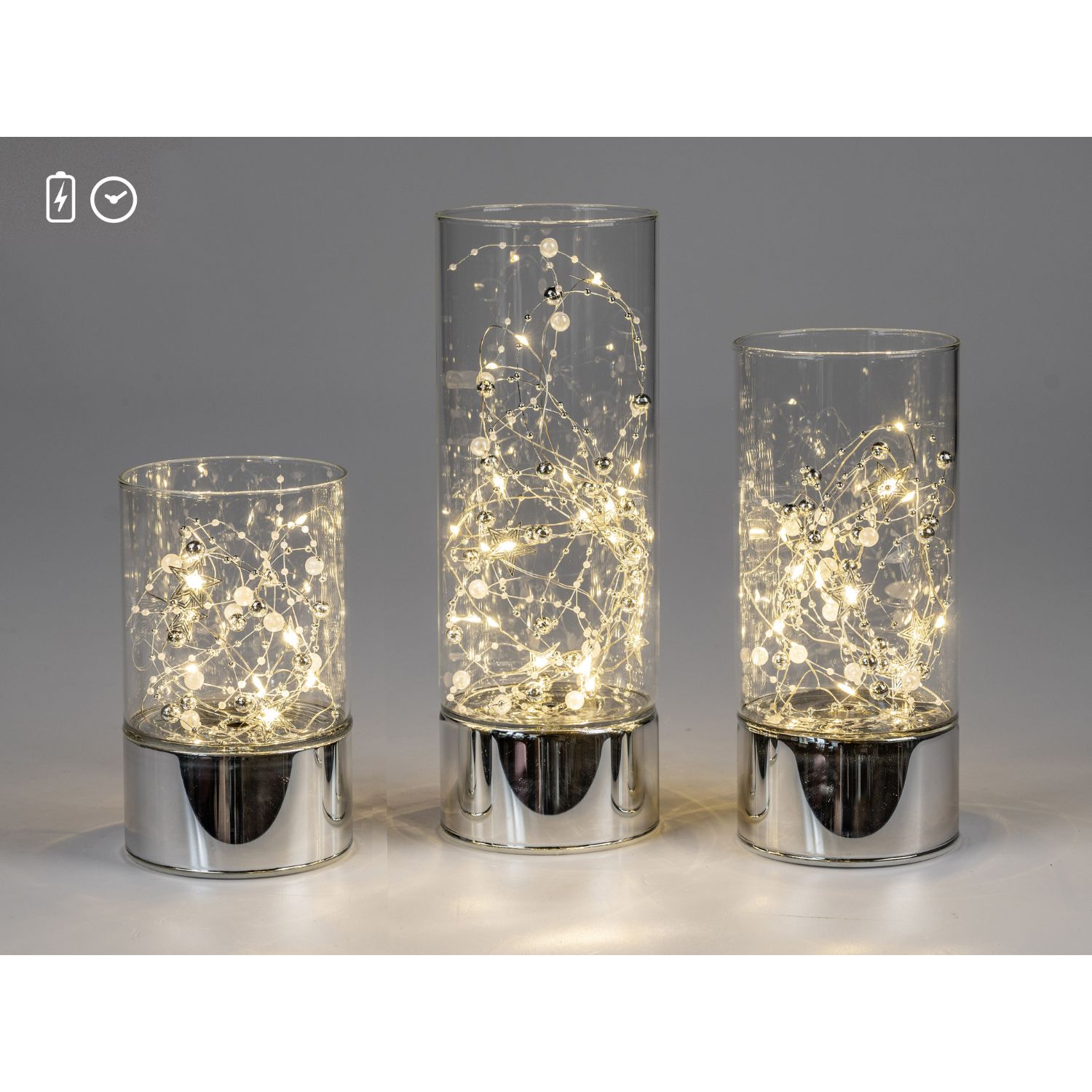 Drei Glaszylinder mit warmweißen LED-Lichterketten auf silbernen Sockeln vor grauem Hintergrund