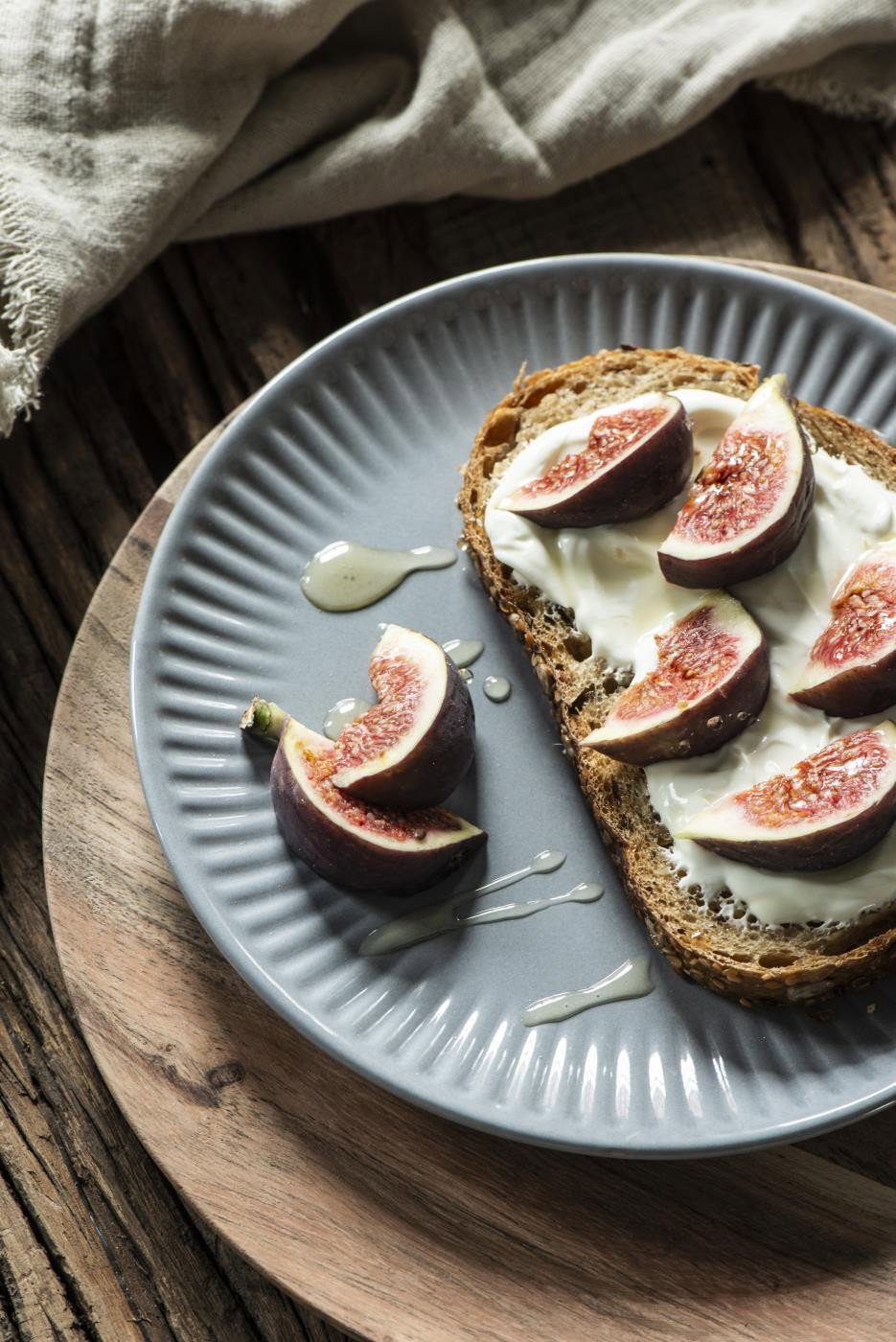Ein grauer Teller mit einem Stück Brot, belegt mit Joghurt und Feigen, daneben ein kleiner Tropfen Honig. Holzuntergrund.