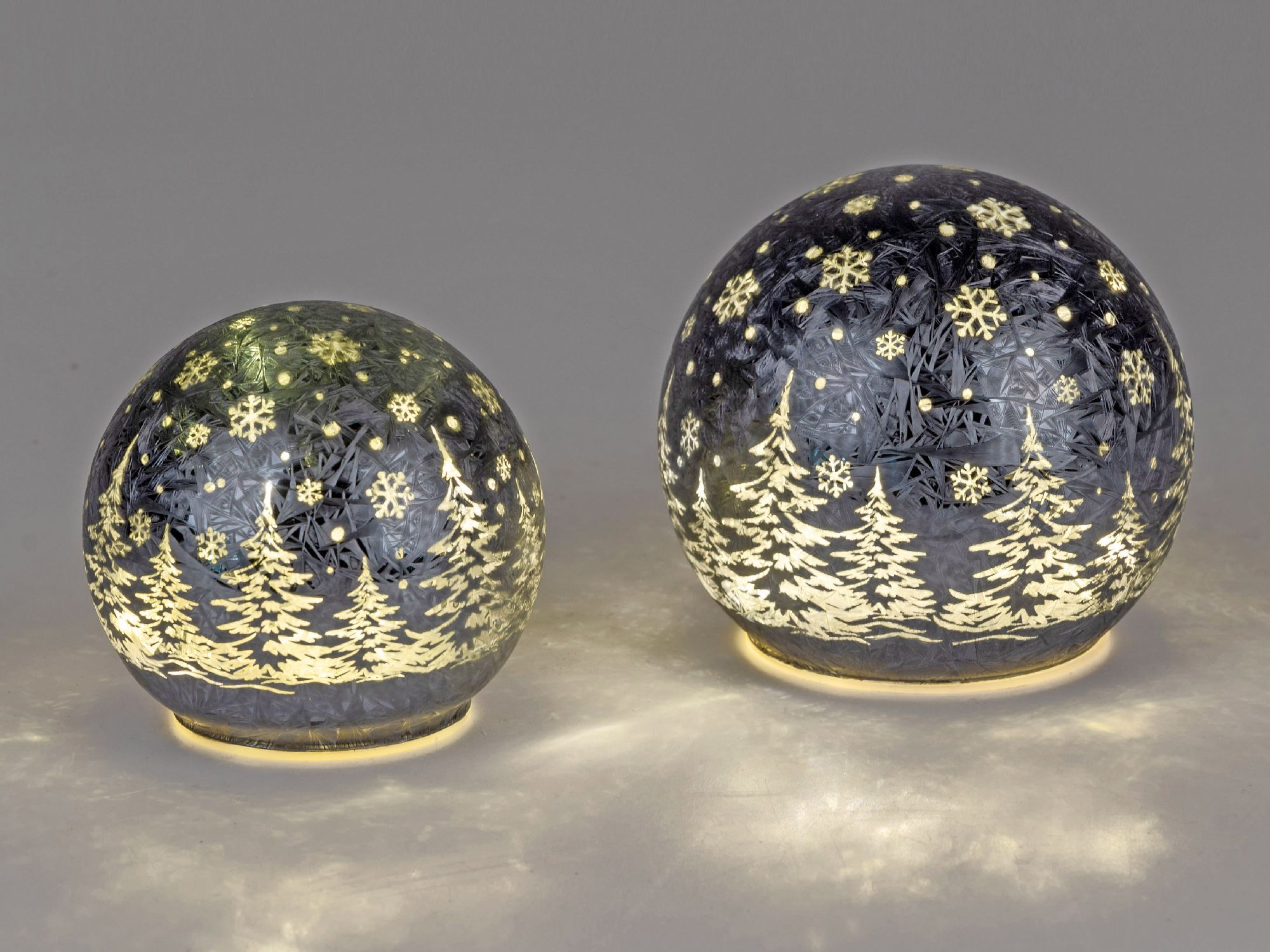 Zwei gläserne Kugeln mit winterlichen Motiven, verziert mit Tannenbäumen und Schneeflocken, beleucht