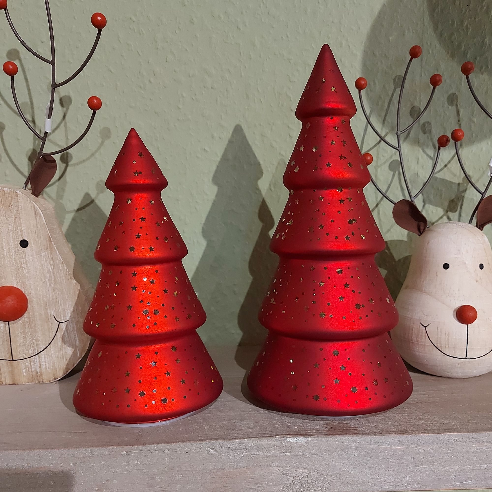 Zwei rote Weihnachtsbaum-Dekorationen aus Keramik stehen auf einem Tisch, umgeben von weiteren festl