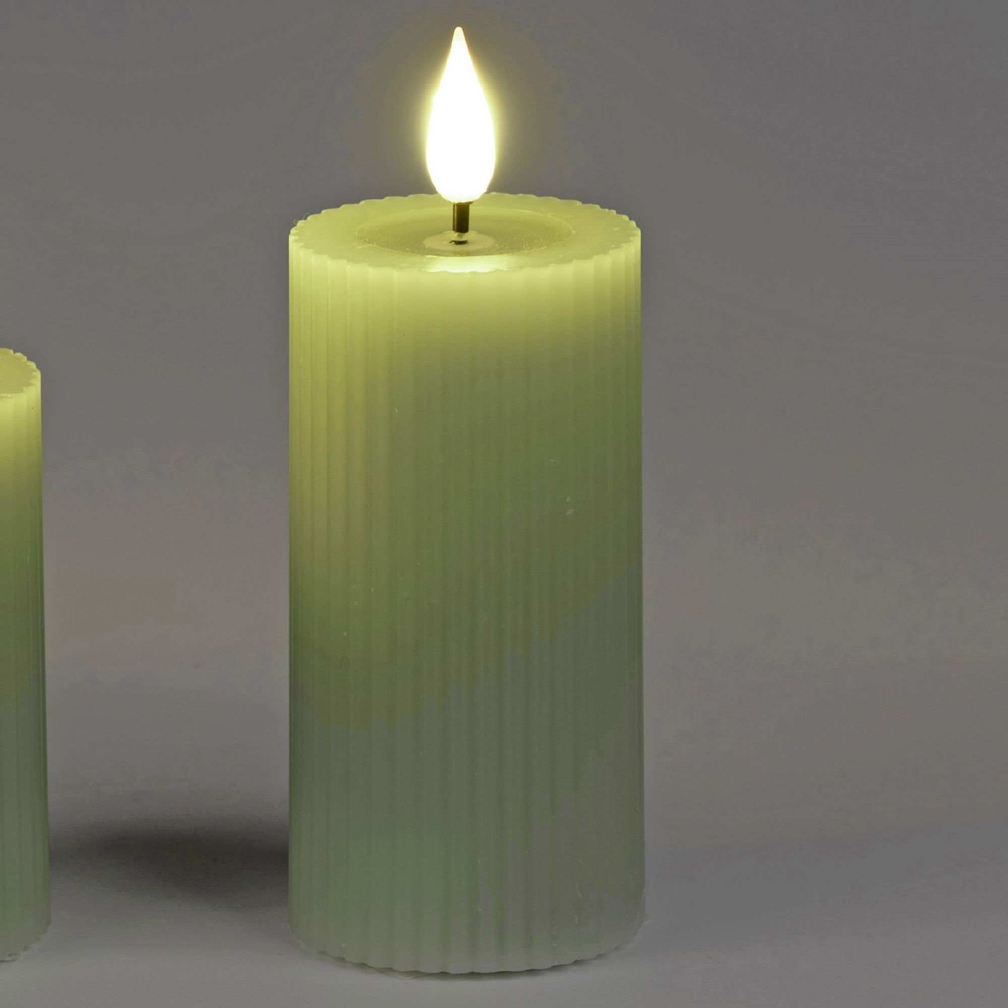 Zwei grüne, gerippte Kerzen stehen nebeneinander. Eine Kerze hat eine flackernde, künstliche Flamme. Hintergrund ist grau.