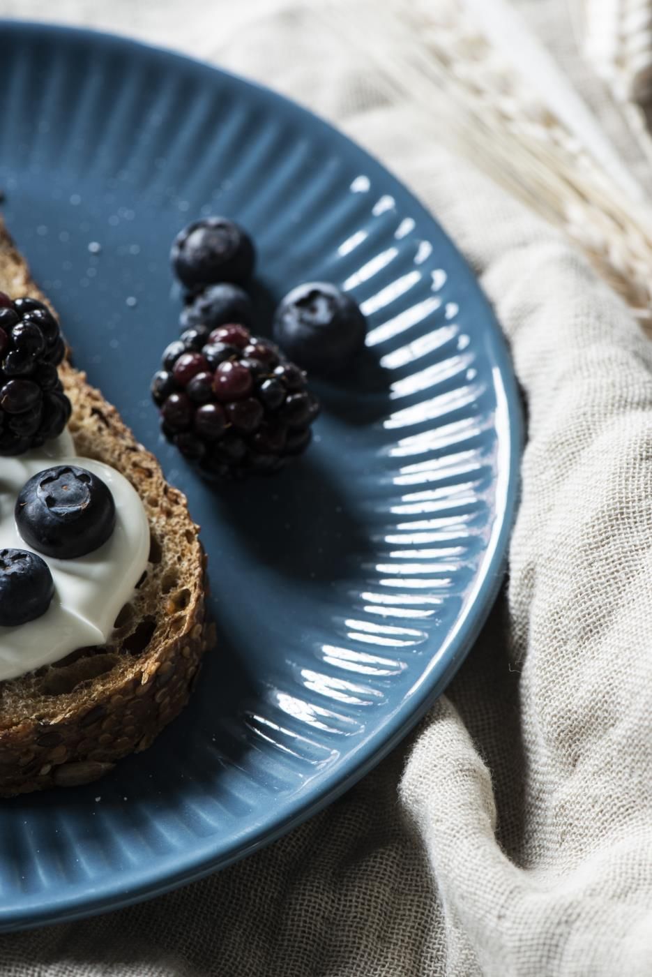 Ein blauer Teller mit einem Stück Vollkornbrot, Joghurt und Heidelbeeren sowie Brombeeren. Im Hintergrund ein heller Stoff.