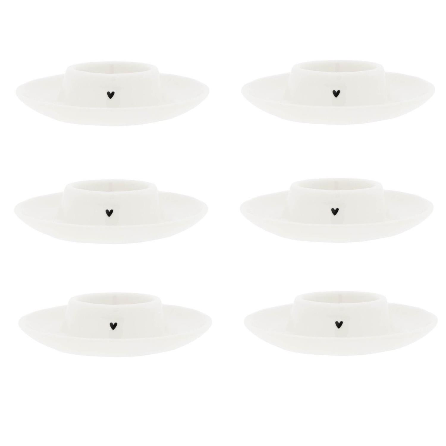 Sechs weiße Kerzenhalter mit einem kleinen schwarzen Herzmotiv auf der Vorderseite, gleichmäßig angeordnet.