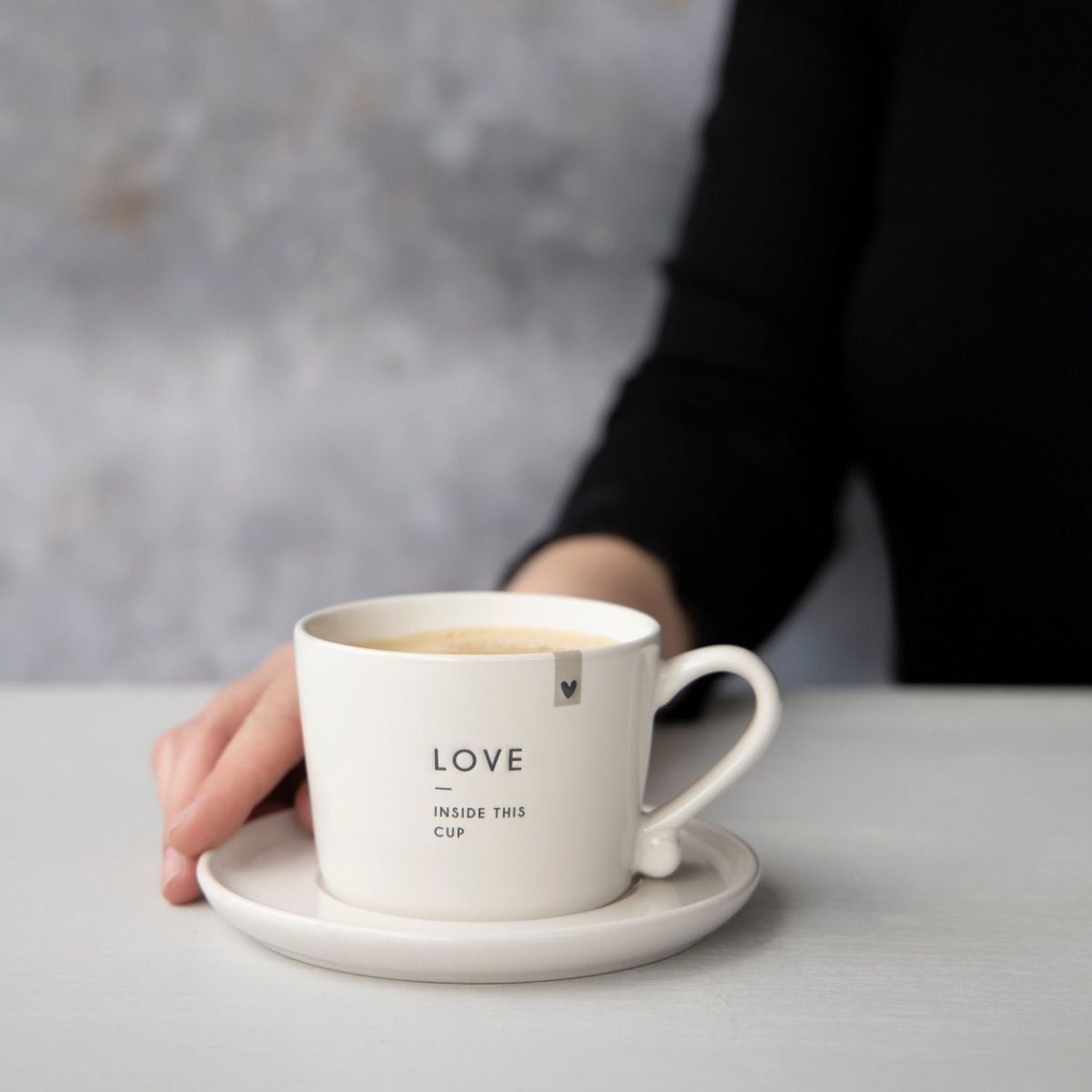 Hand neben weißer Tasse mit Heißgetränk auf Untertasse, grauer Hintergrund