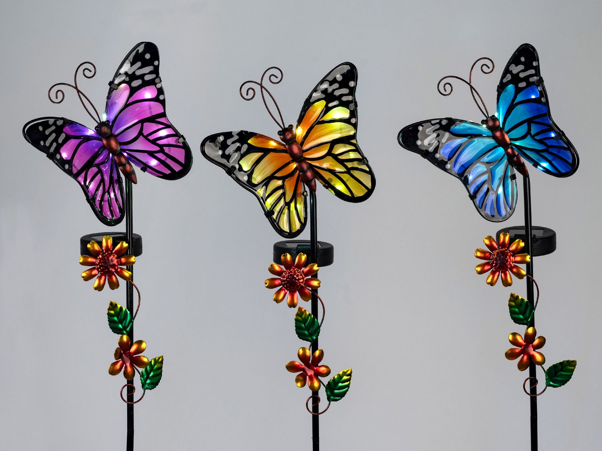 Drei bunte Schmetterlinge aus Glas auf Stäben, umgeben von Blumen und Blättern, vor einem hellen Hin