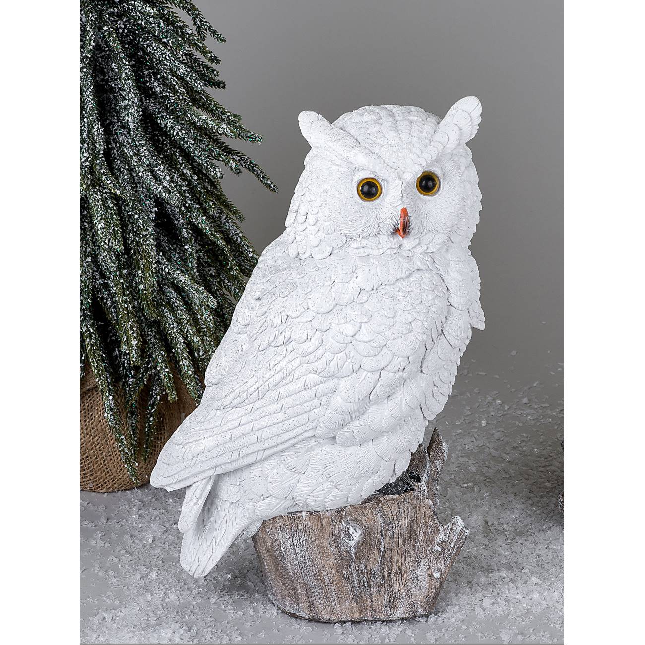 Eine weiße Eule sitzt auf einem Holzstamm, umgeben von einer grünen Pflanze und einer schneebedeckte