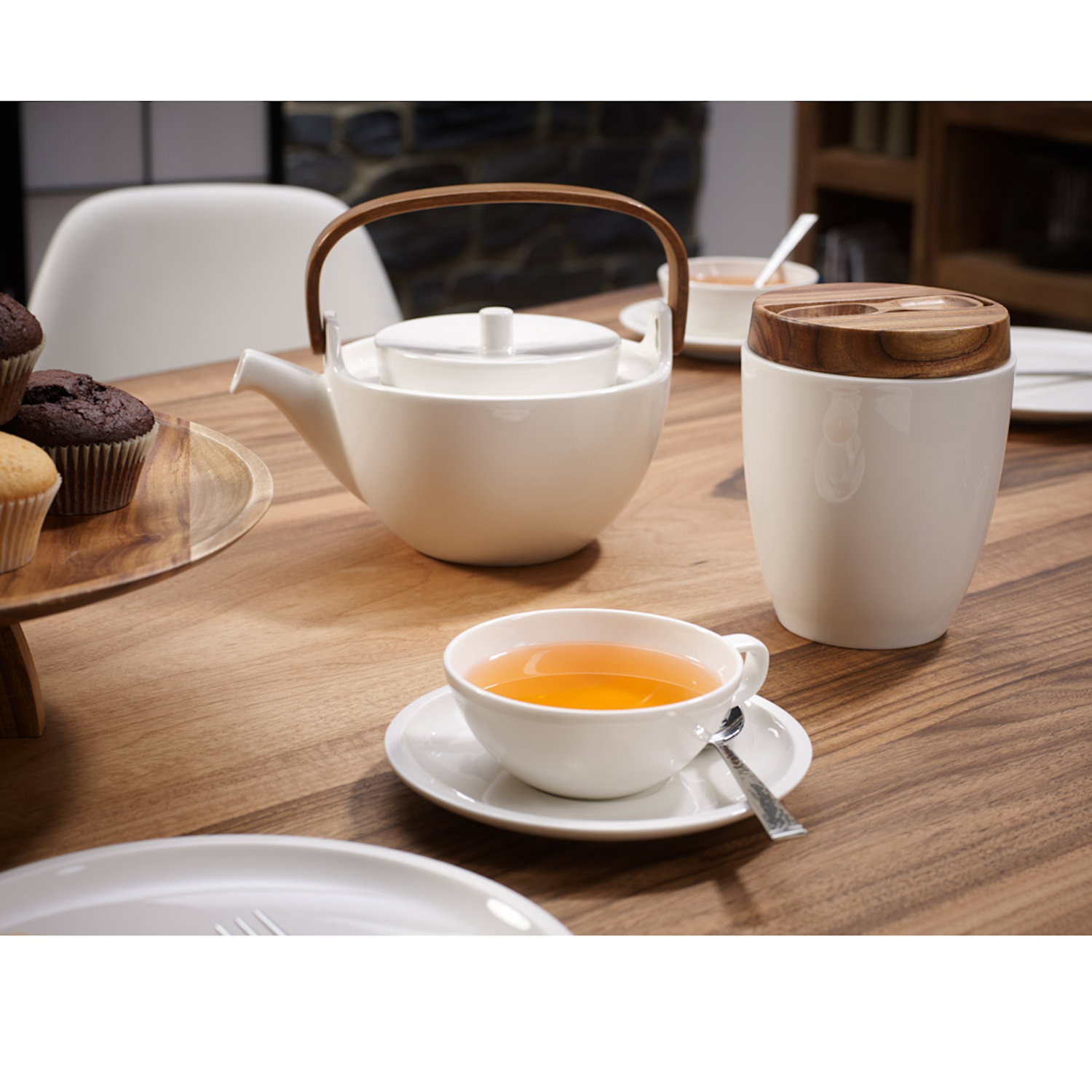 Ein Tisch mit einer weißen Teekanne, einer Tasse mit Tee, einem Becher mit Holzdeckel und Muffins auf einem Holztablett.