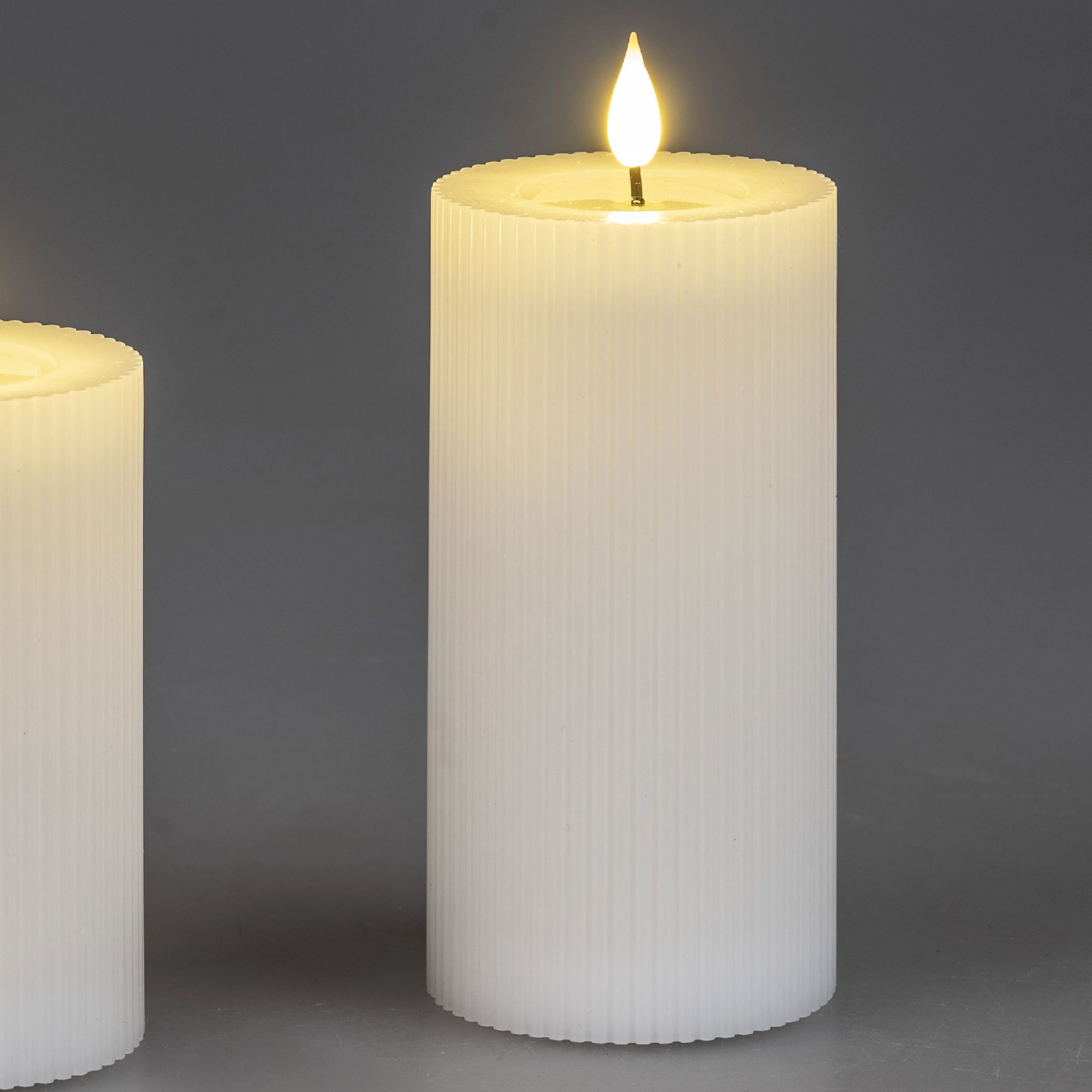 Zwei weiße, gerippte Kerzen stehen vor einem grauen Hintergrund. Eine Kerze brennt, die Flamme leuchtet sanft.