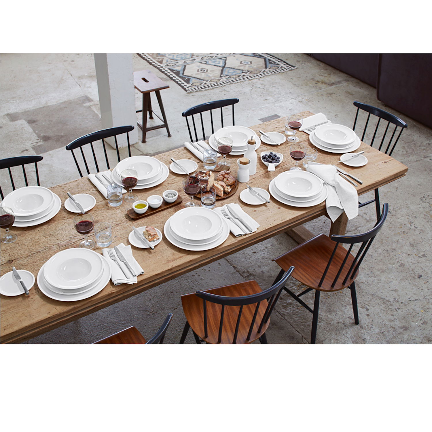 Ein gedeckter Holztisch mit weißen Tellern, Gläsern, Besteck und verschiedenen Speisen. Stühle mit schwarzem Gestell und Holzsitz.