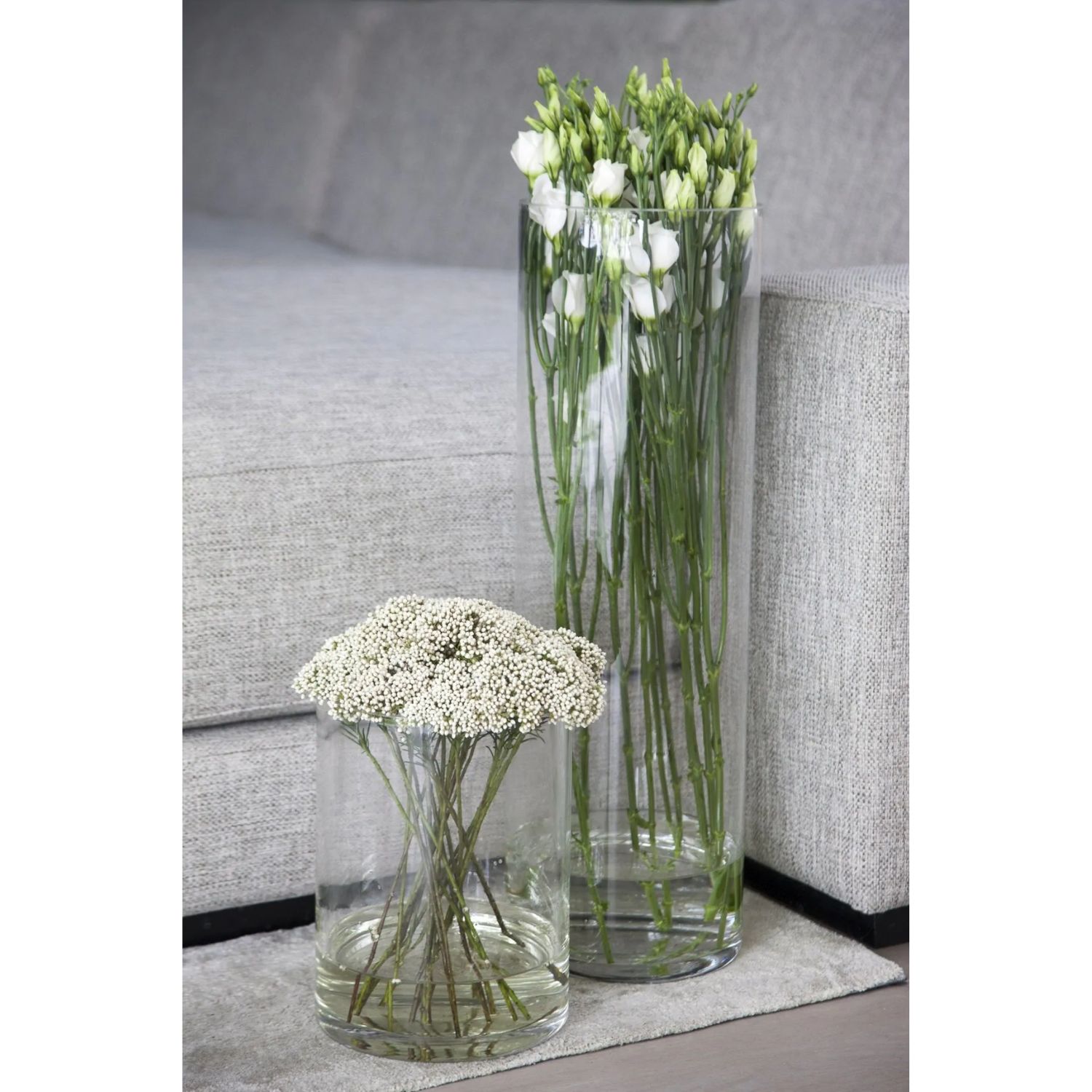 Zwei Glasvasen mit Blumen: Eine hohe Vase mit weißen Rosen und eine niedrigere mit kleinen weißen Blüten. Im Hintergrund eine graue Couch.