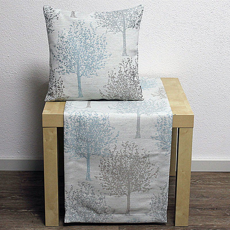 Ein Kissen und ein Tischläufer mit Baum-Muster in Blau- und Grautönen auf einem hellen Tisch. Hintergrund in neutralem Farbton.