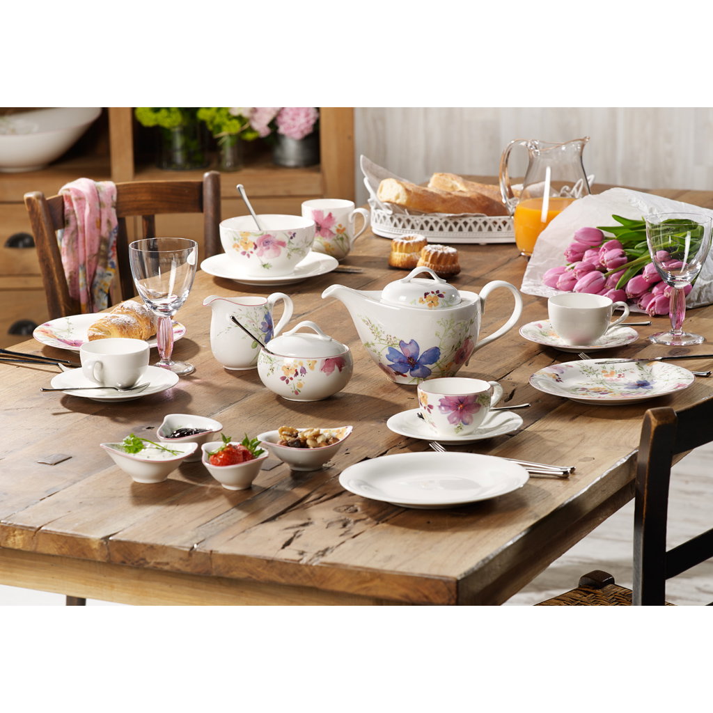 Ein gedeckter Tisch mit Geschirr, Teekanne, Gläsern, Brötchen, Obst und Blumen in einer gemütlichen Umgebung.