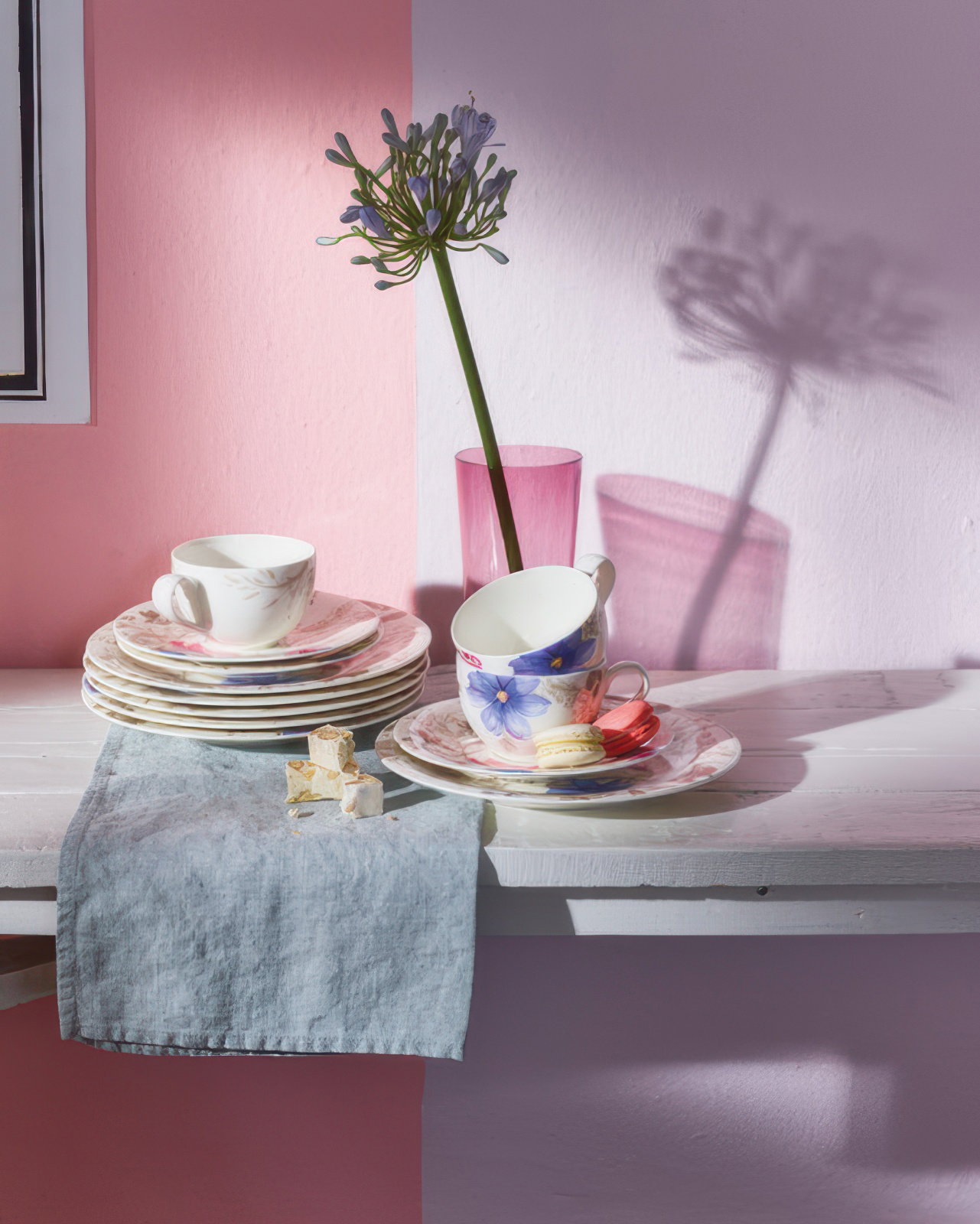 Teller, Tassen und ein Blumenstrauß stehen auf einem Tisch mit einer blauen Serviette. Die Wand ist rosa gestrichen.