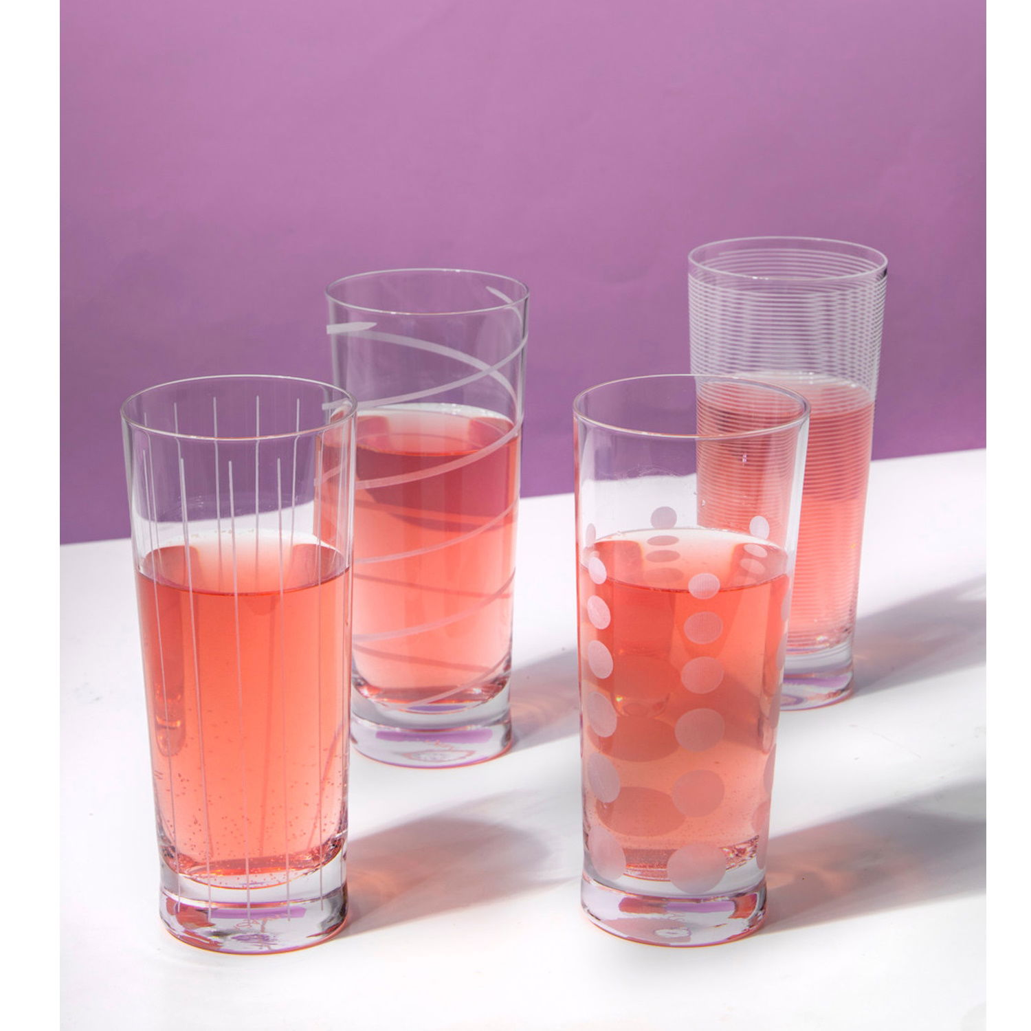 Vier Gläser mit pinkem Getränk stehen auf einem weißen Tisch vor einem lila Hintergrund. Jedes Glas hat ein unterschiedliches Muster.