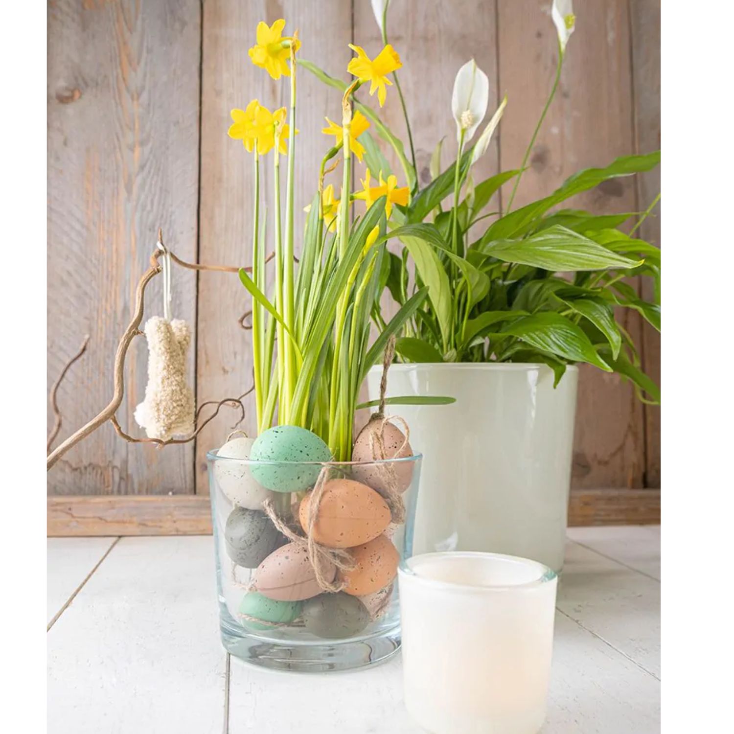 Blumentopf mit gelben Narzissen in Glasvase mit bunten Eiern, daneben grüne Zimmerpflanze und Kerze