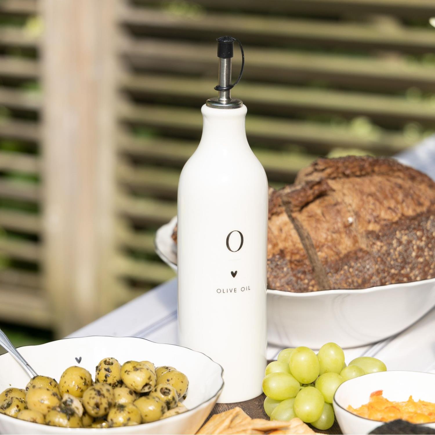Eine weiße Olivenölflasche steht neben einer Schüssel mit grünen Oliven, Trauben und einem Laib Brot
