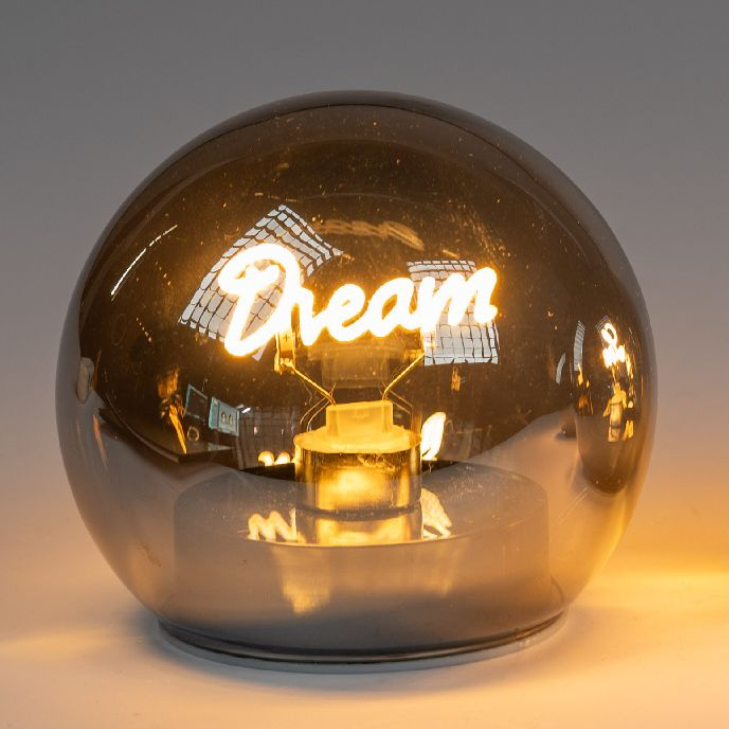 Eine runde, gläserne Lampe mit dem Schriftzug "Dream" in warmem Licht, auf einem hellen Untergrund.