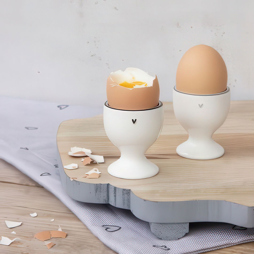 Zwei Eier in weißen Eierbechern auf einem Holzbrett, eines mit zerbrochener Schale und gelbem Eigelb, das andere unversehrt.