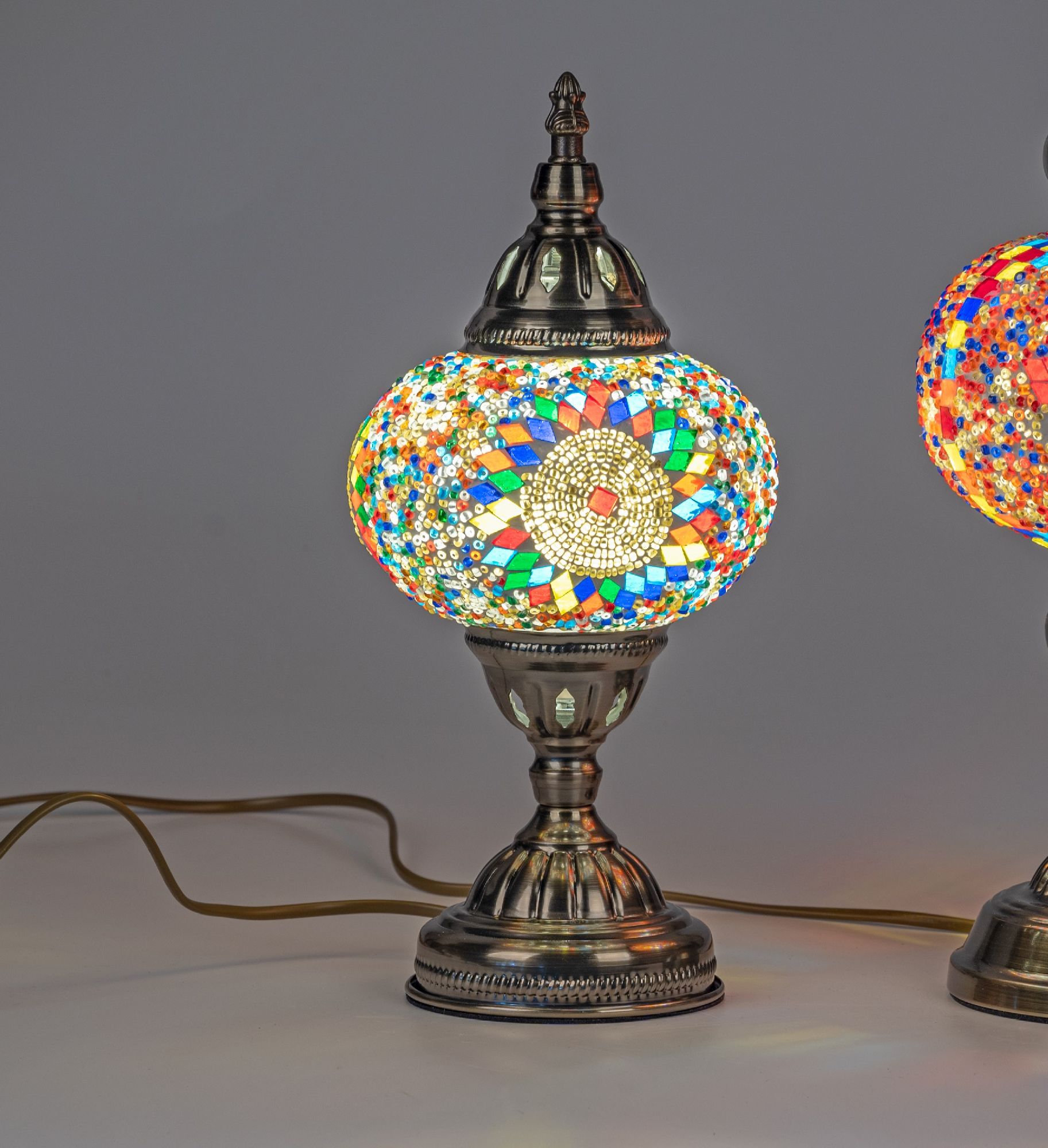 Bunte Tischlampe mit mosaikartigem Glasdesign und metallischem Fuß, auf grauem Hintergrund. Kabel si