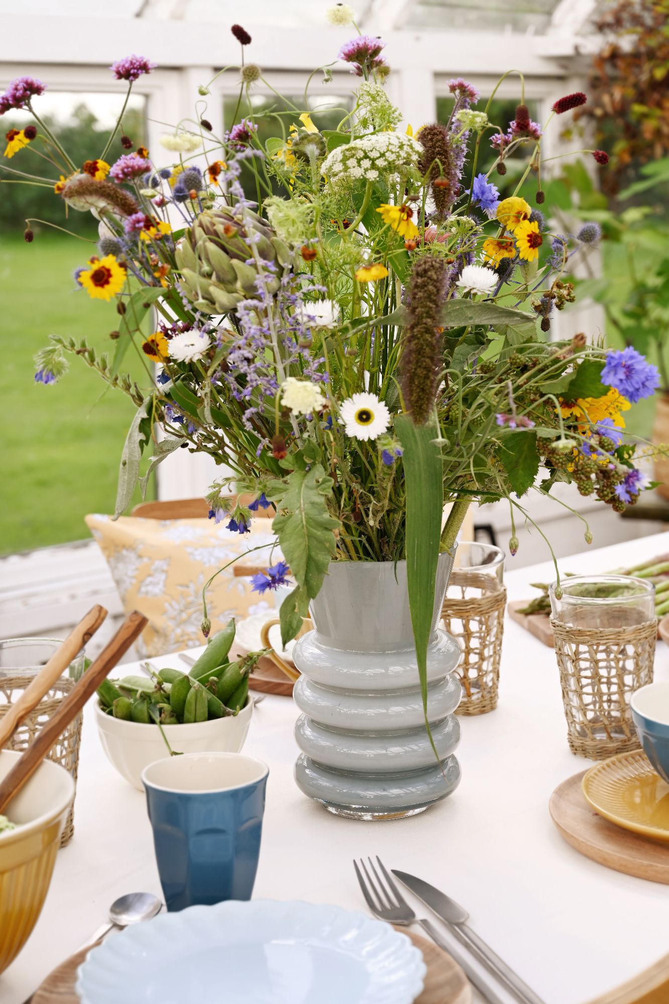 Ein Tisch mit einem bunten Blumenstrauß in einer Vase, Geschirr, Besteck und Schalen mit Lebensmitteln. Hintergrund mit Fenster.