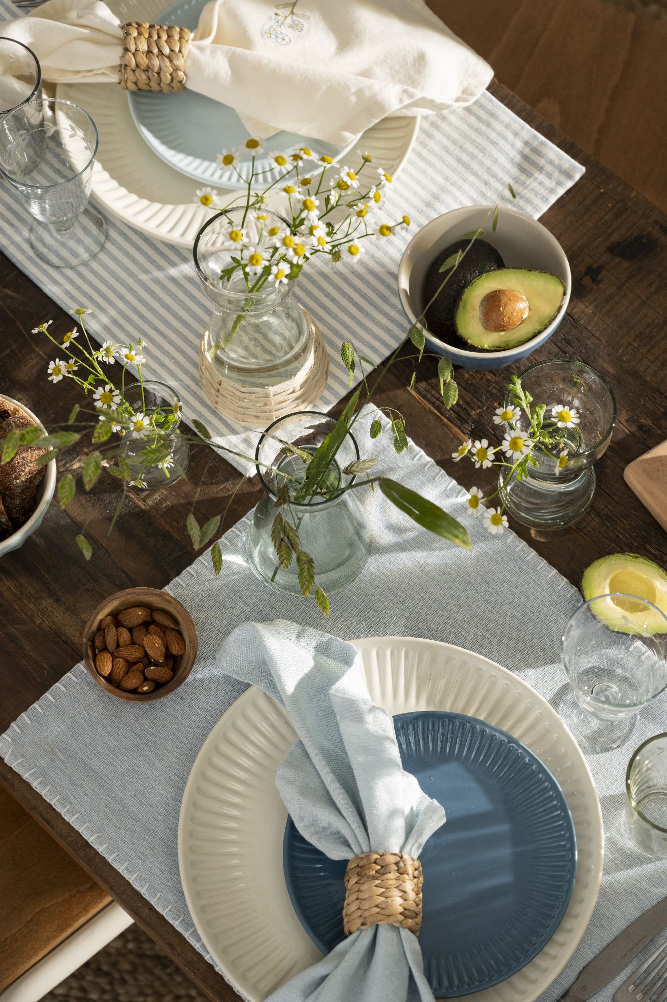 Tischgedeck mit Tellern, Gläsern, einer Schüssel mit Avocado, Blumen in Vasen und einer Schale mit Mandeln auf einem Tisch.