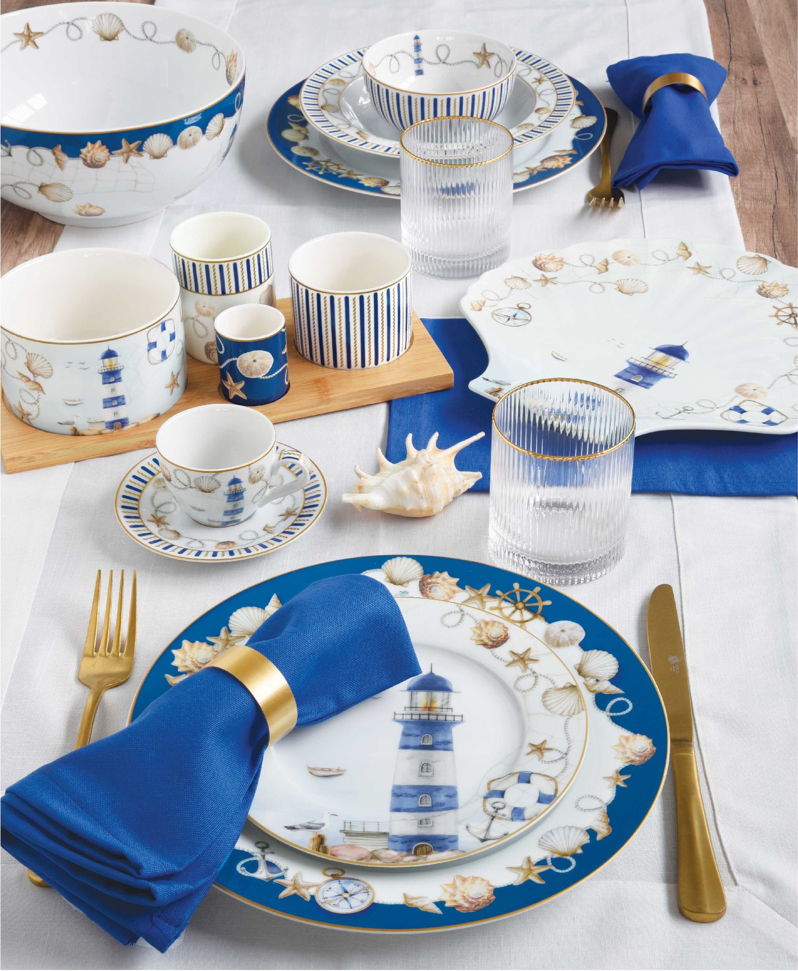 Tischgedeck mit blau-weißer Porzellanserie, Besteck, Gläsern und Servietten auf einem weißen Tisch. Dekorative Muscheln sind sichtbar.