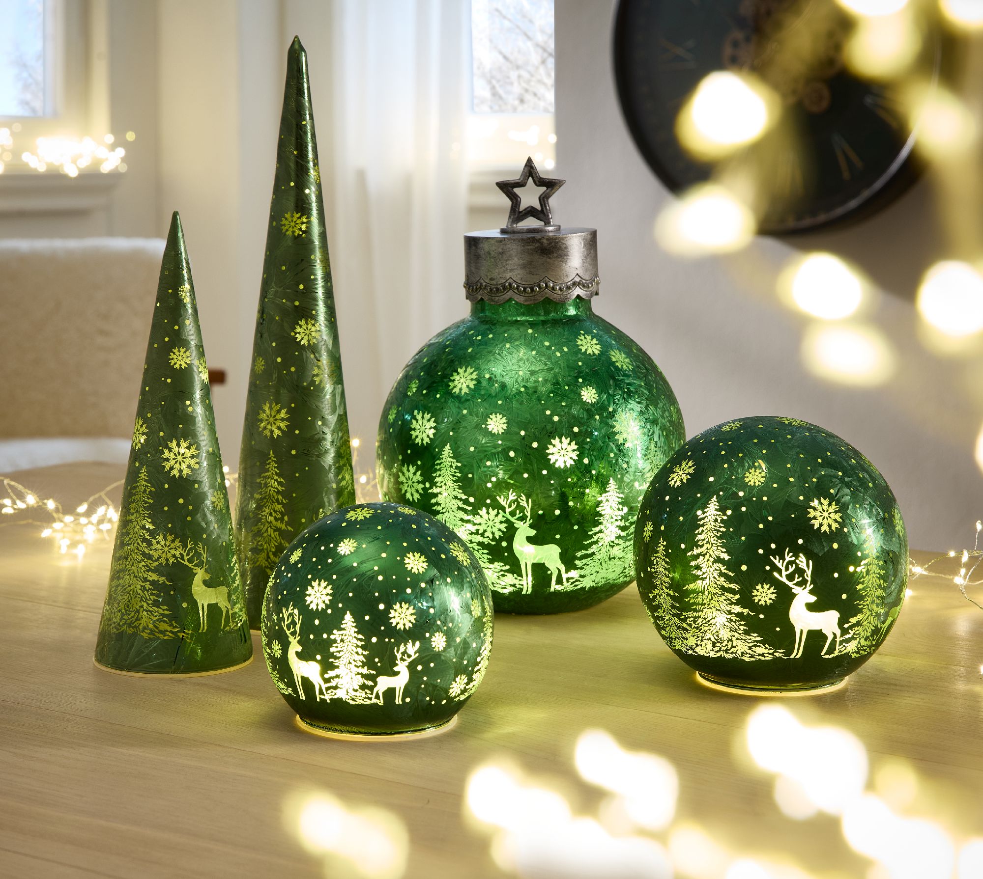 Grüne Dekorationsobjekte in Form von Bäumen und Kugeln mit winterlichen Motiven stehen auf einem Tis