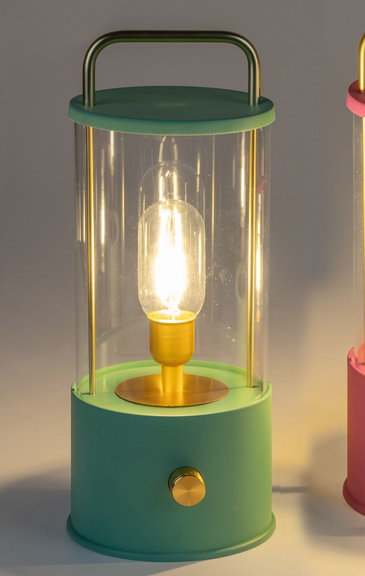 Eine grüne Tischlampe mit transparentem Glas und goldenem Sockel, die leuchtet. Rechts steht eine pi
