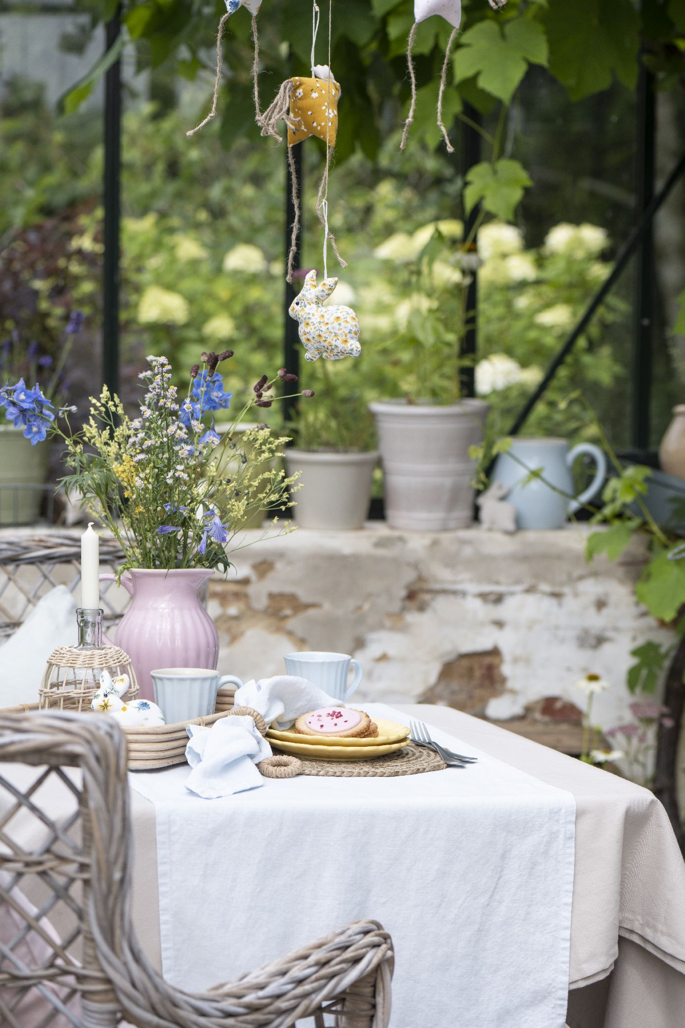 Ein gedeckter Tisch im Freien mit Blumen, Geschirr, Keksen und dekorativen Hängedekorationen. Im Hintergrund Pflanzen und Töpfe.