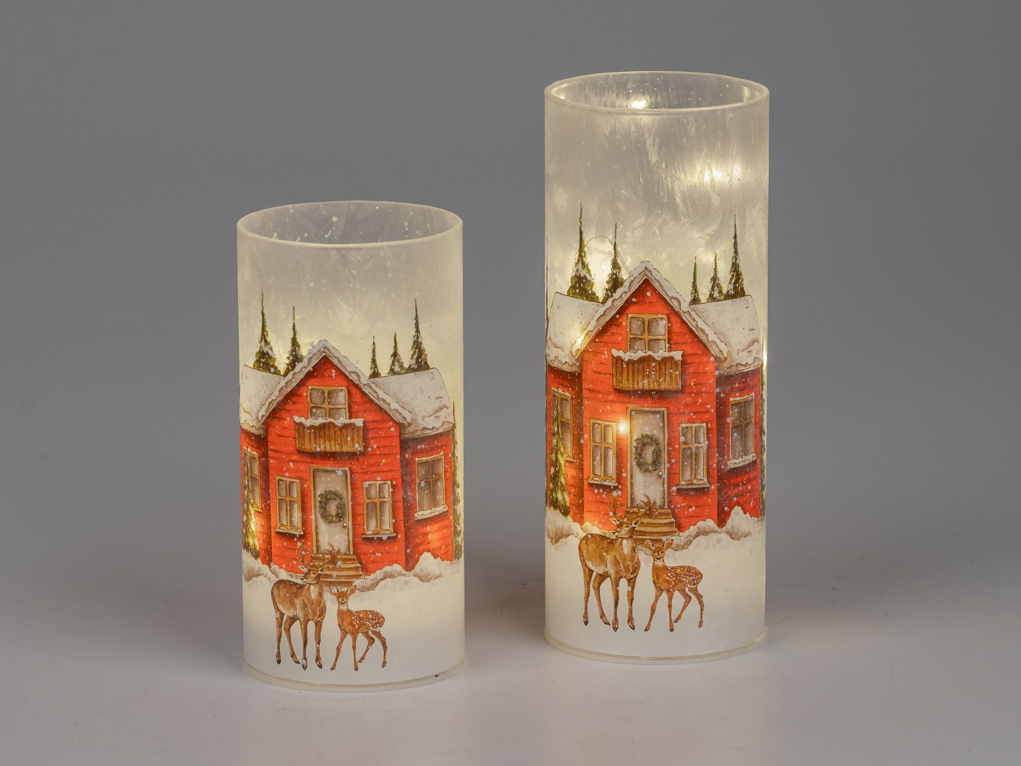 Zwei leuchtende Kerzen mit winterlichem Motiv: ein rotes Haus, Tannenbäume und Rehe im Schnee. Hinte