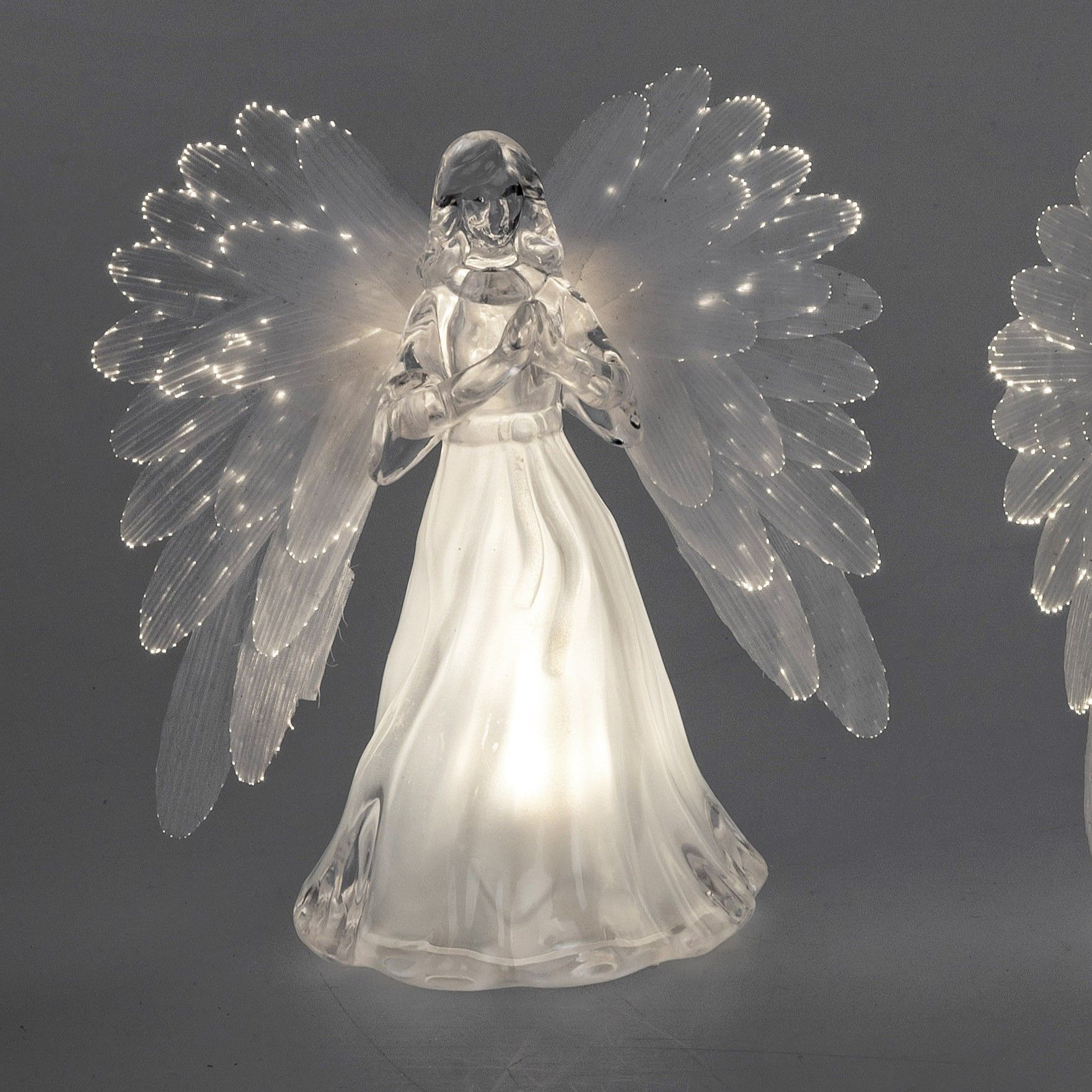 Glasengel mit transparenten Flügeln, in der Mitte beleuchtet, steht vor grauem Hintergrund.