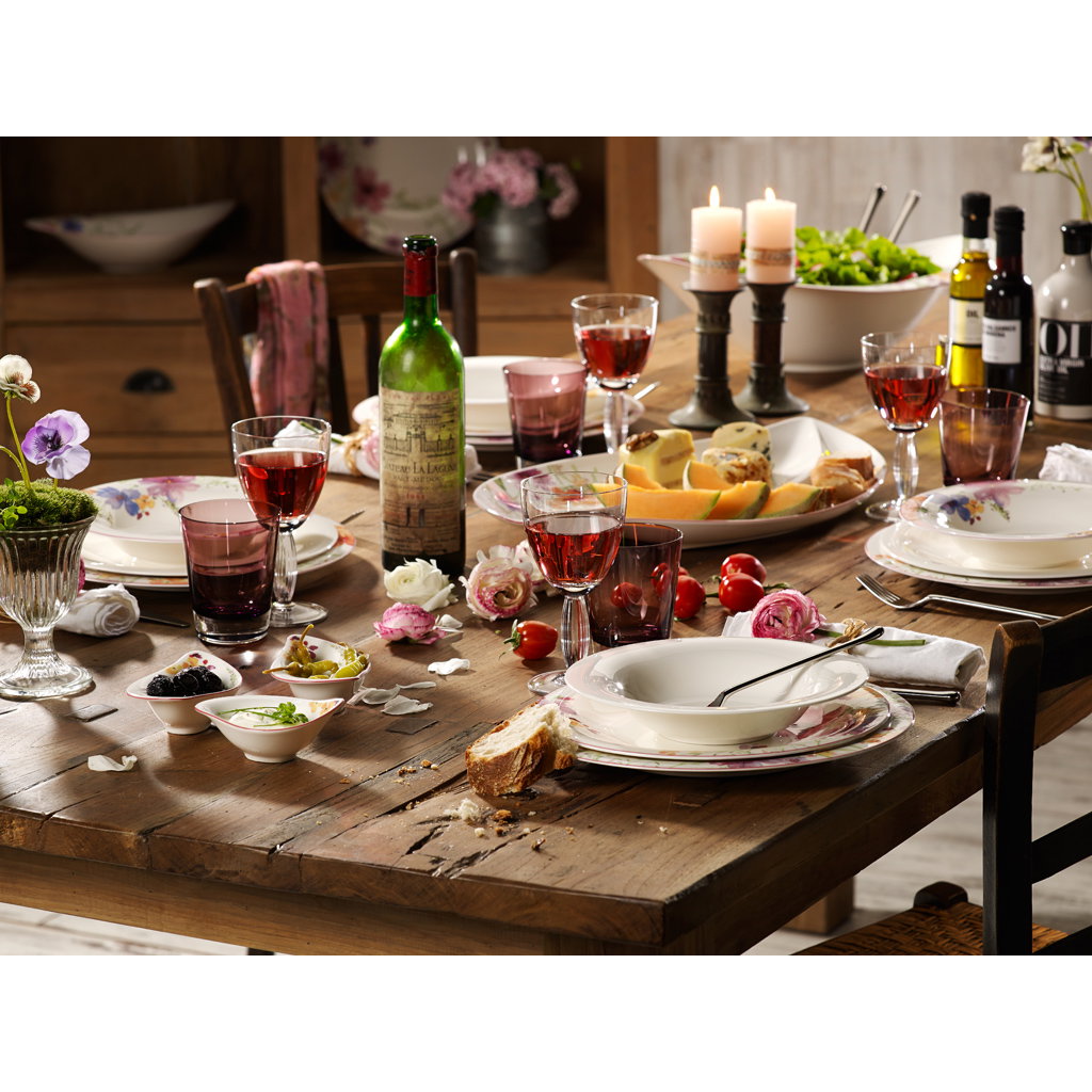 Ein gedeckter Tisch mit Tellern, Gläsern, Weinflasche, Obst, Käse und Blumen. Kerzen und verschiedene Dips sind ebenfalls vorhanden.
